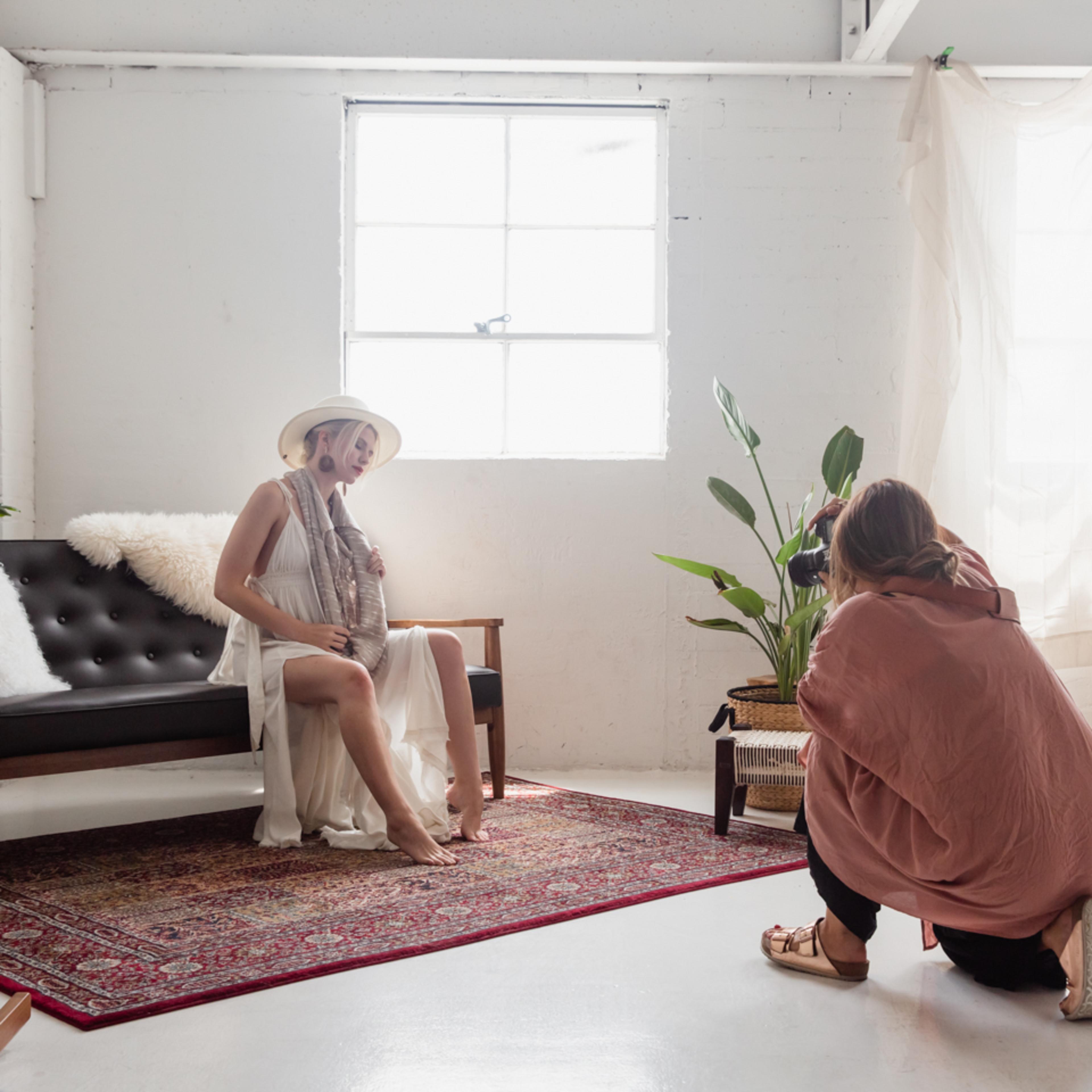 A photographer captures a model sitting on a couch in a bright, minimalist studio with large windows and indoor plants.