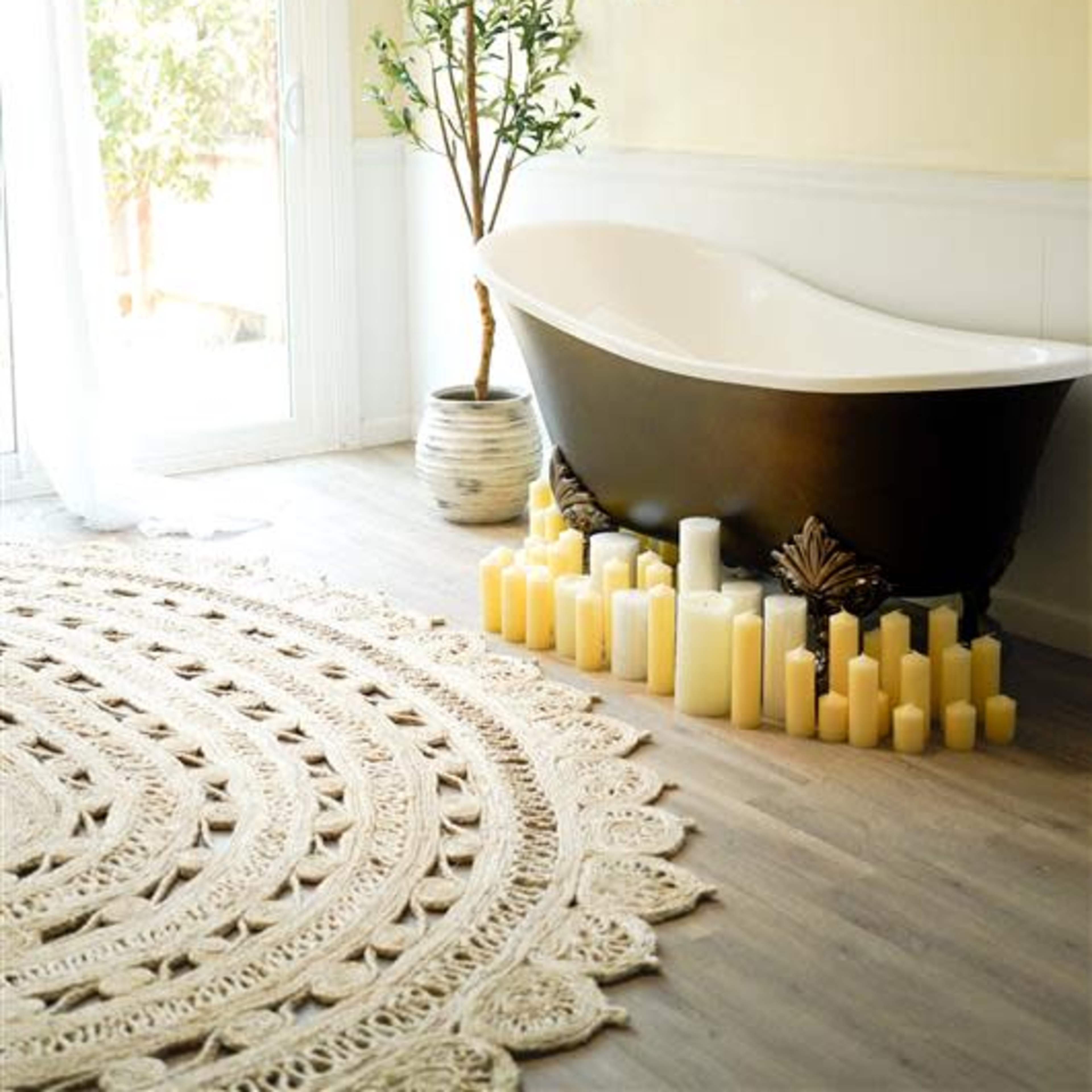 A freestanding black bathtub is positioned beside a large window, surrounded by a circular decorative rug and multiple cream-colored candles.