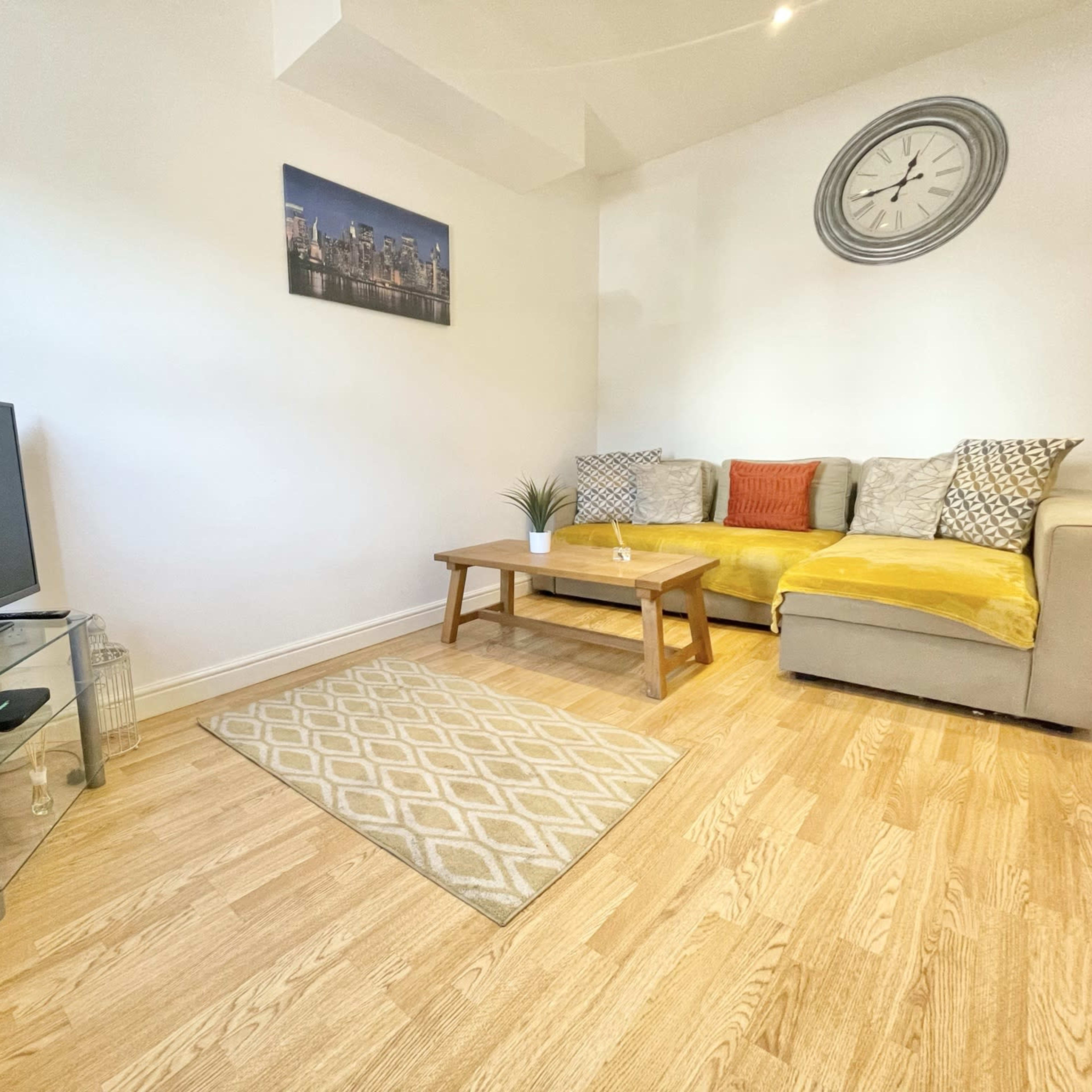 A living room with a corner sofa, a coffee table on a patterned rug, a television on a glass stand, and a wall clock above.
