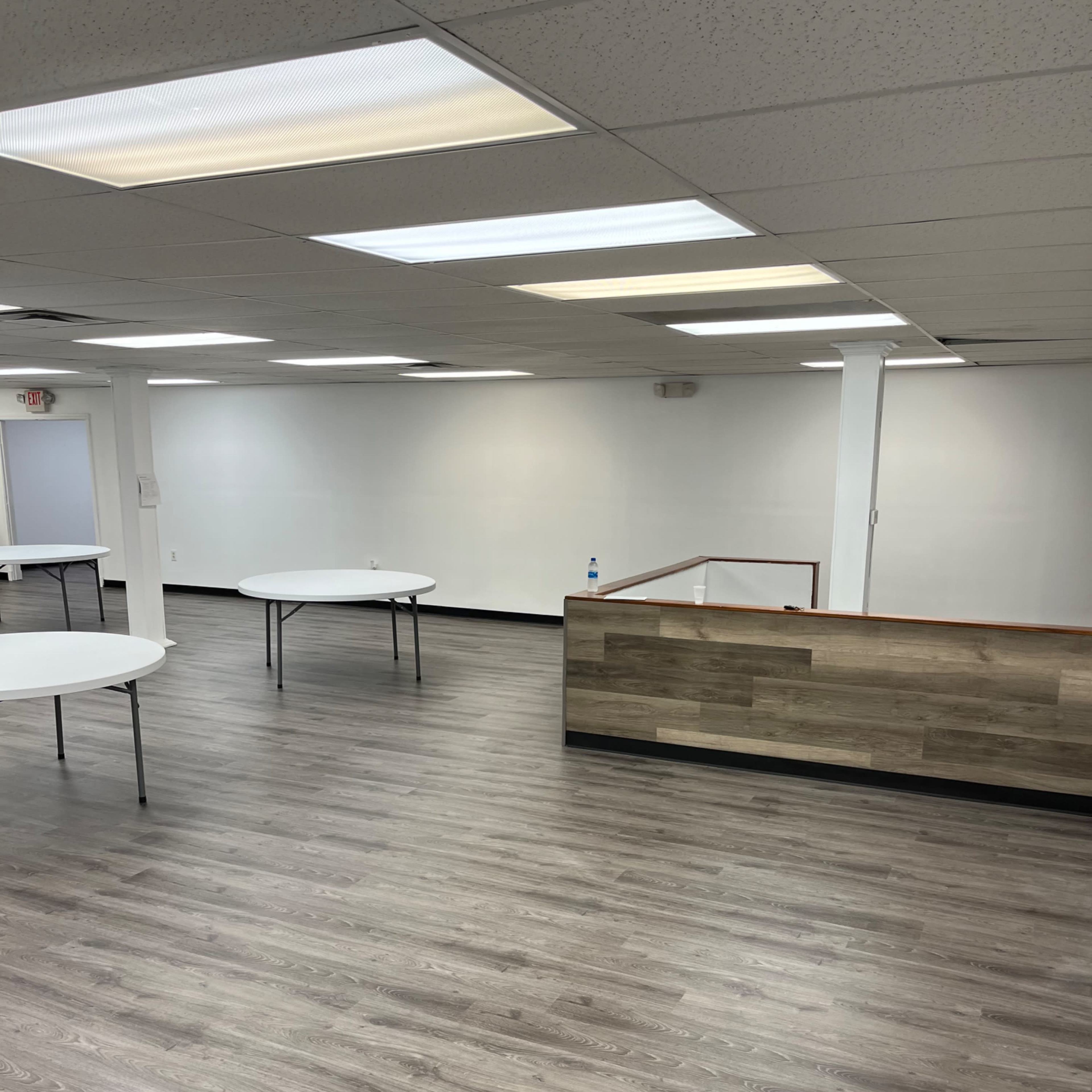 An empty room with light-colored walls, several round tables, and a wooden reception area in the background.