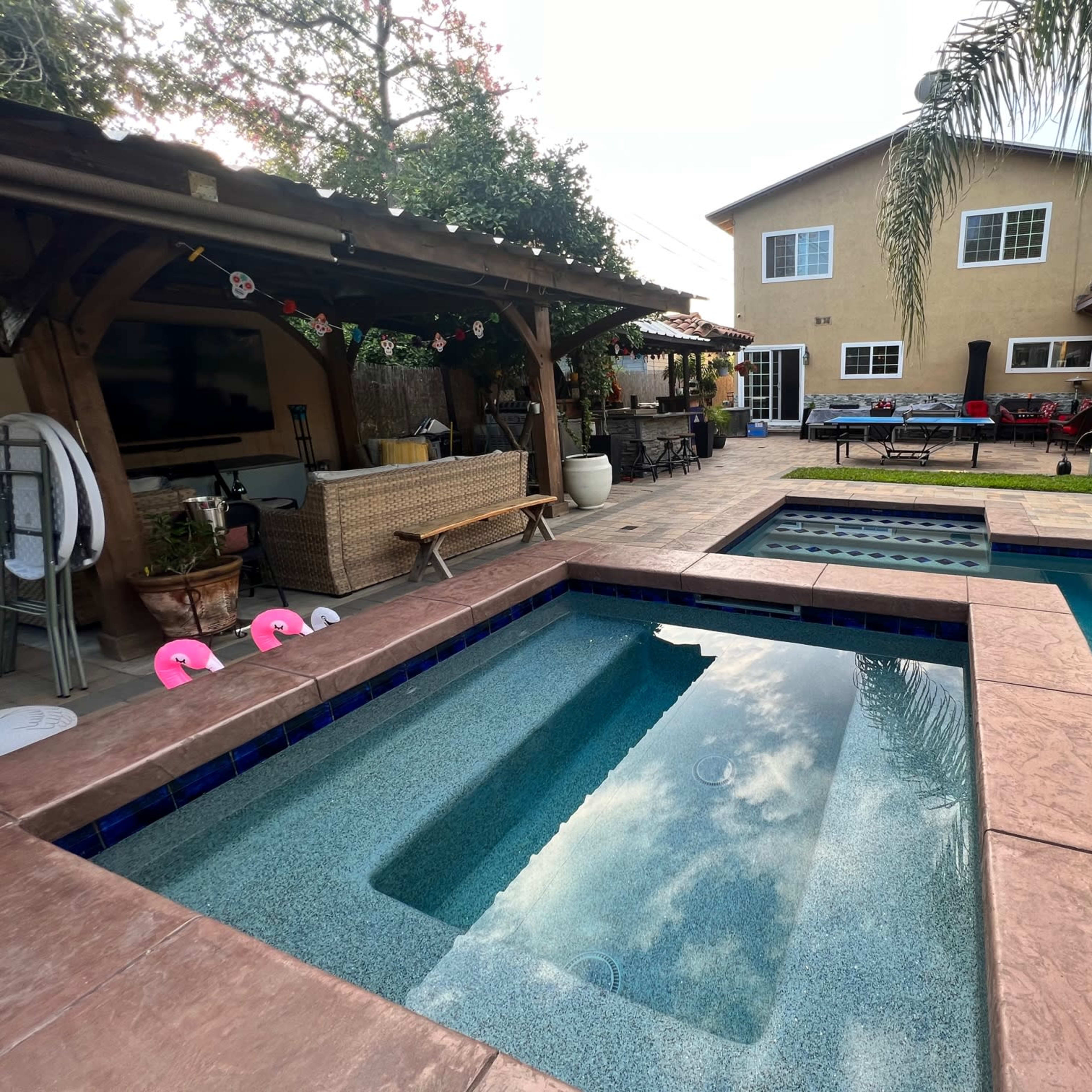 A backyard features a swimming pool with a spa, surrounded by concrete decking and a two-story house in the background.