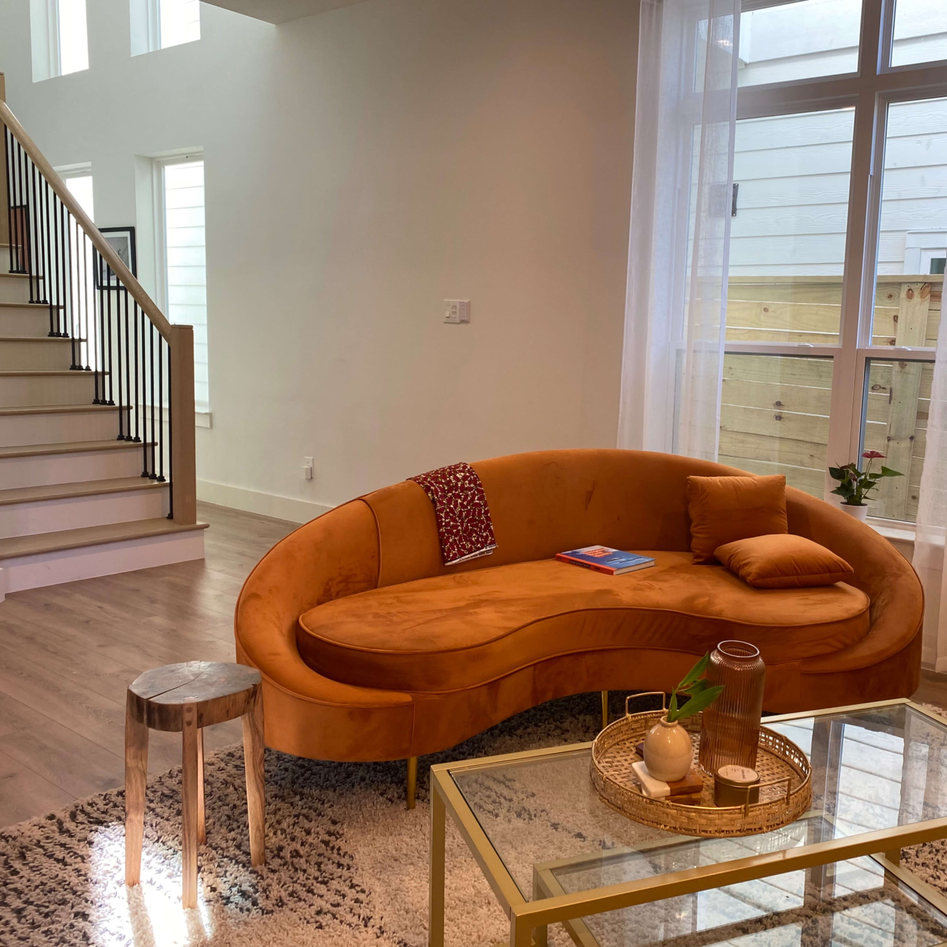 A curved orange sofa is positioned next to a glass coffee table and a small wooden stool in a bright, modern living room with large windows.