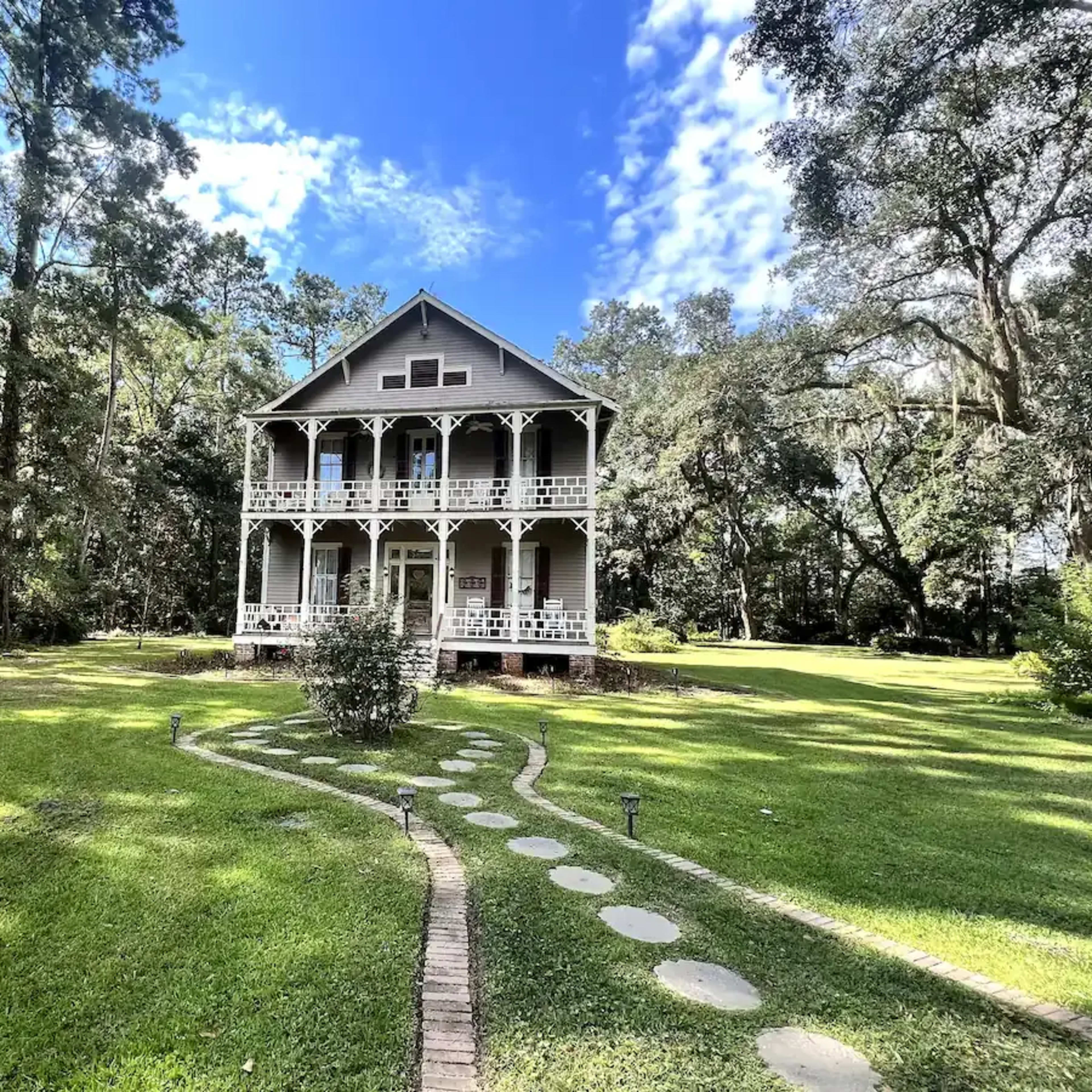 A two-story house with a wraparound porch is set in a landscaped yard surrounded by trees.