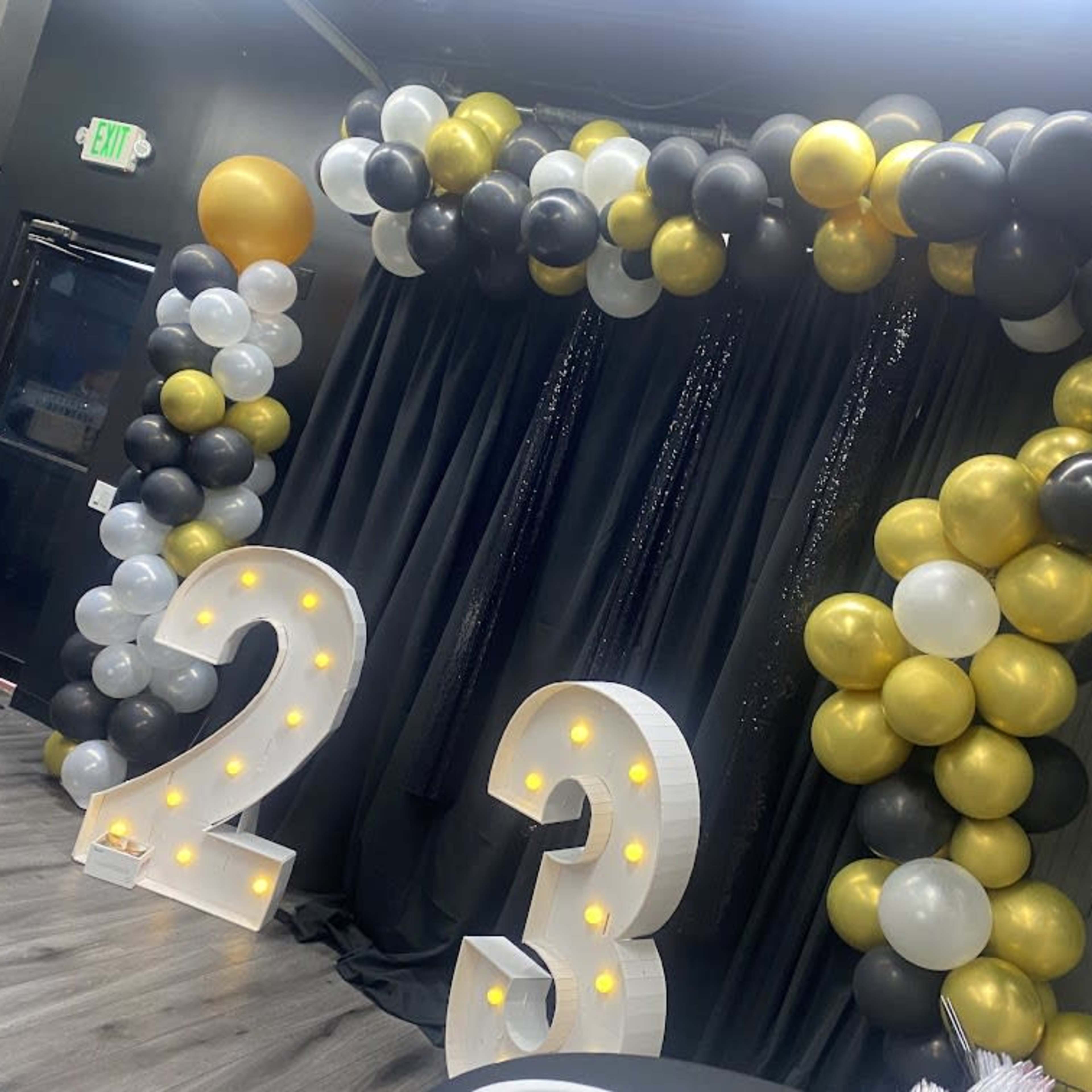 A decorative backdrop featuring a balloon arch in gold, black, and white colors, with large illuminated numbers "2" and "3" in front of a black curtain.