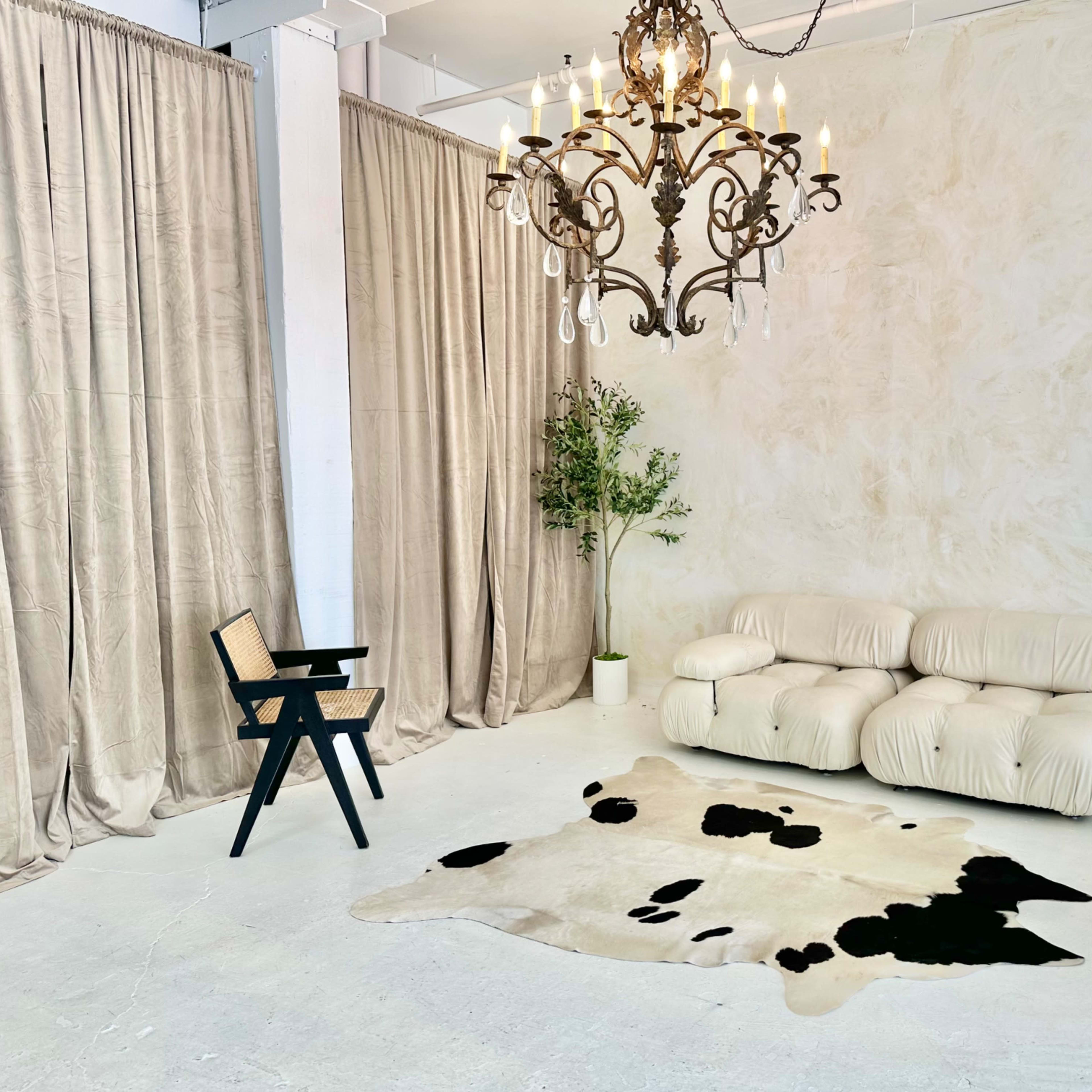 A minimalist interior space featuring a beige wall, a plush cream-colored sofa, a black and white cowhide rug, a dark wooden chair, and a chandelier overhead.