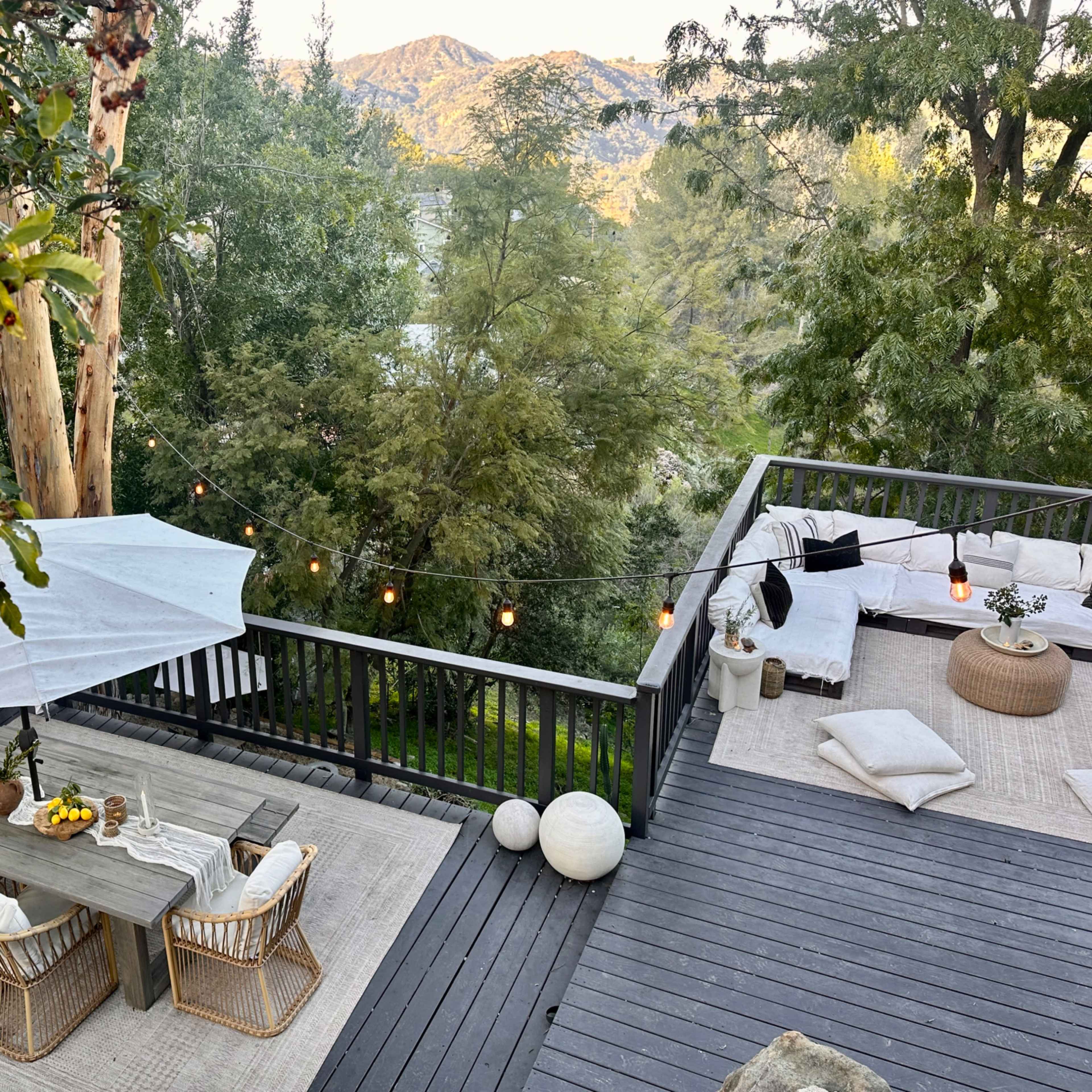 A wooden deck features a dining table with chairs, surrounded by greenery and mountain views, illuminated by string lights.