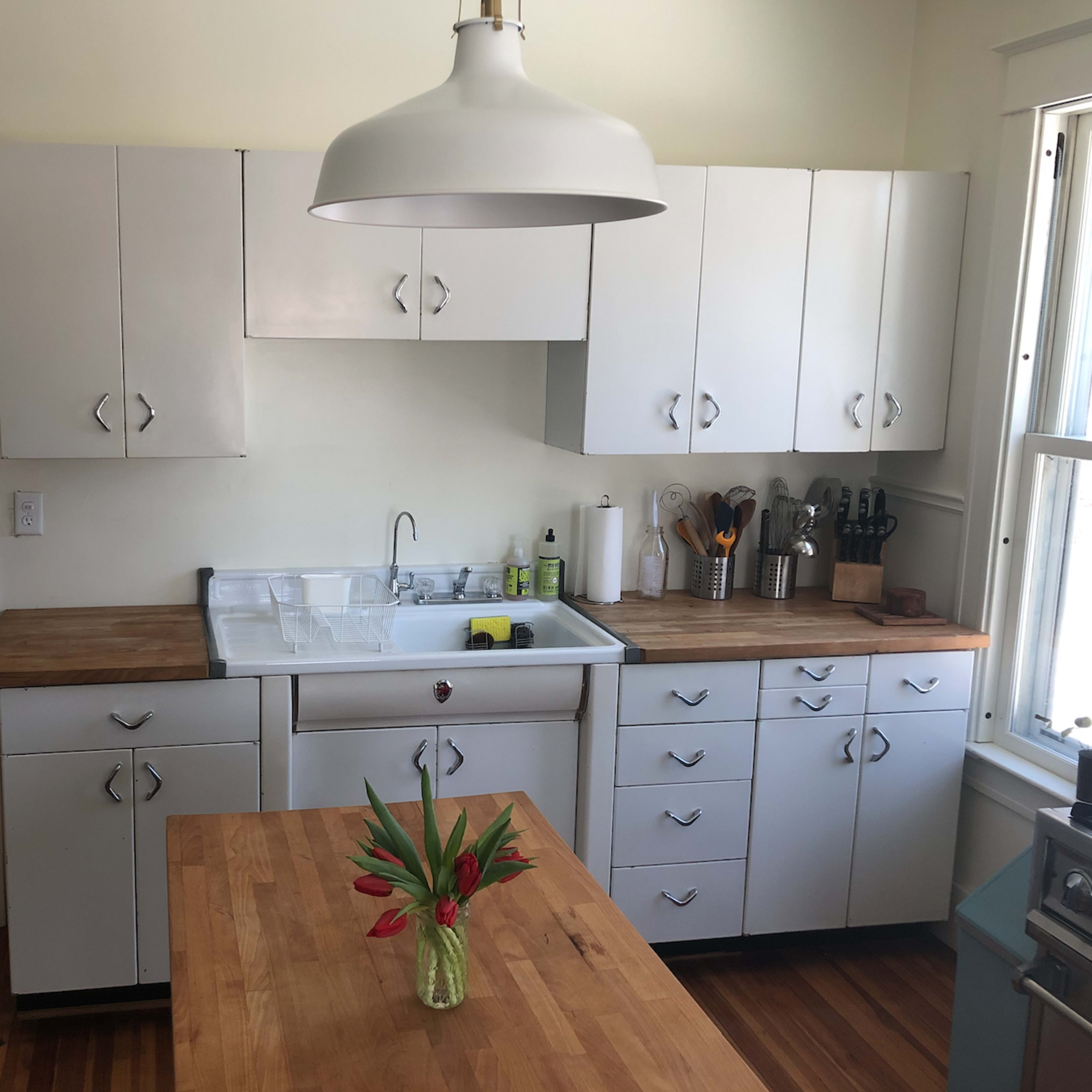A clean kitchen features white cabinets, a wooden countertop, and a sink with a window above it, complemented by a vase of flowers on the table.