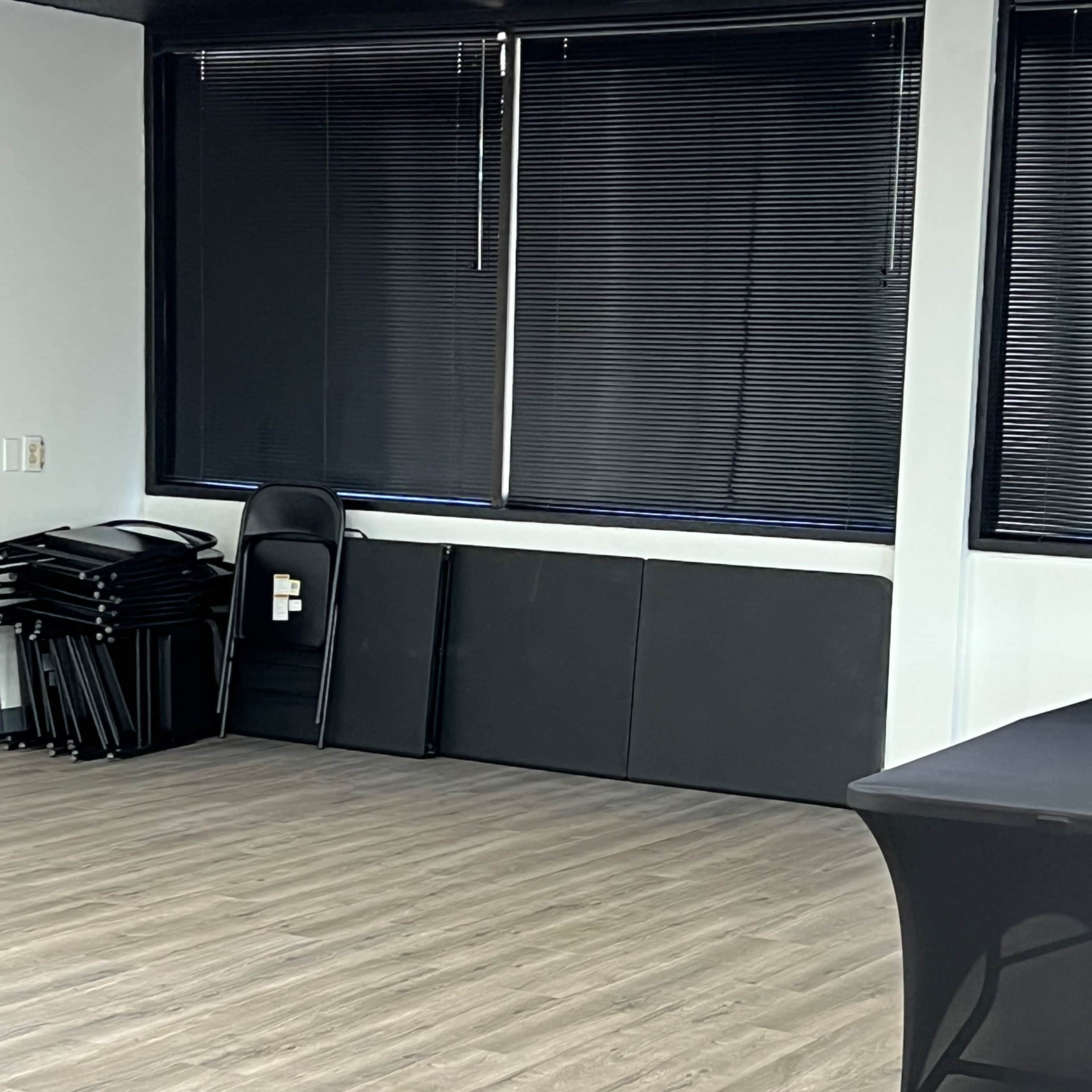 An empty room with a few stacked chairs and black folding mats against the wall, alongside a table covered with a black cloth.