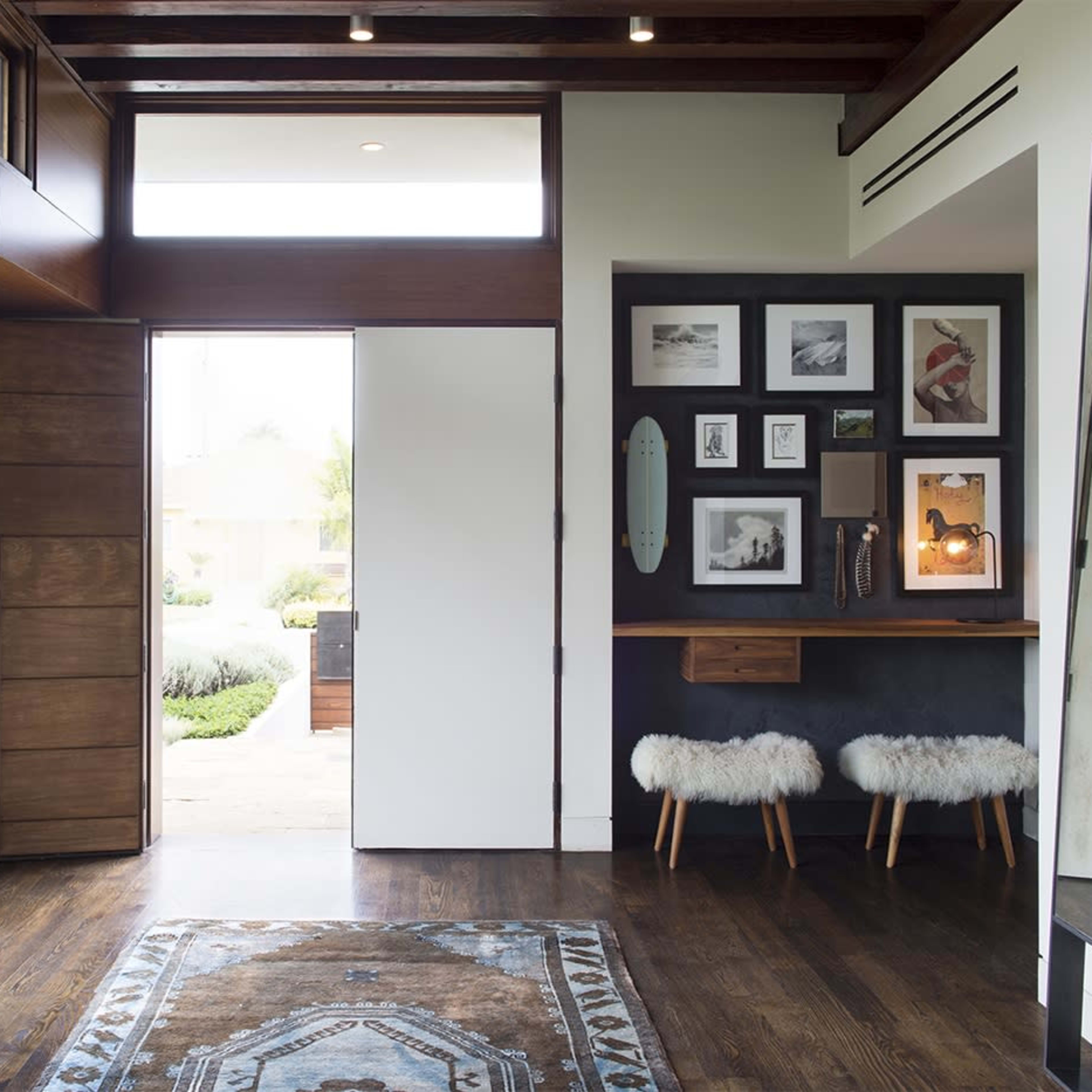A modern entryway featuring a large window, a door leading outside, a small table with a decorative arrangement, and a wall adorned with framed pictures next to two upholstered stools.