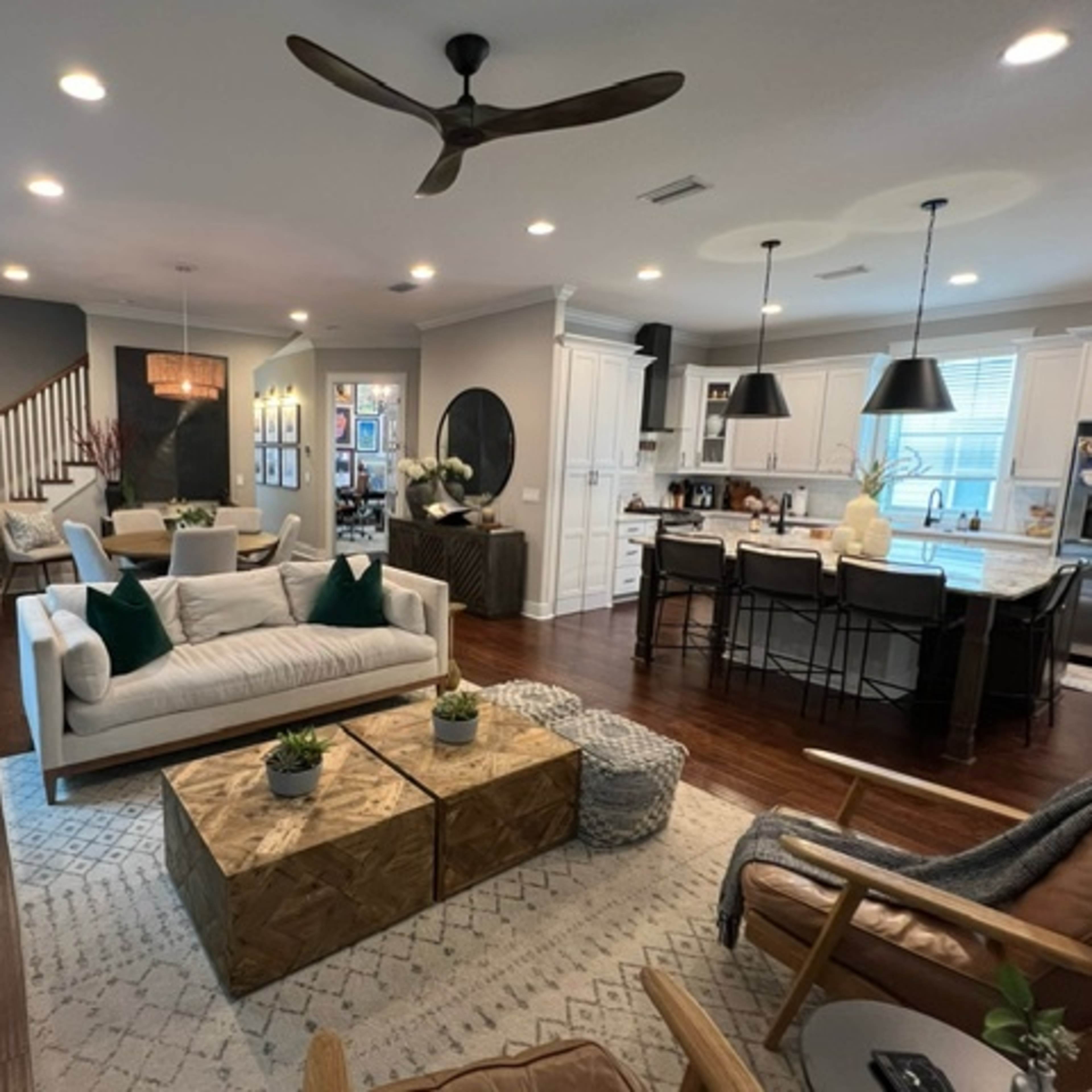 A modern living room with a sectional sofa, two wooden coffee tables, and a kitchen in the background featuring white cabinetry and bar seating.