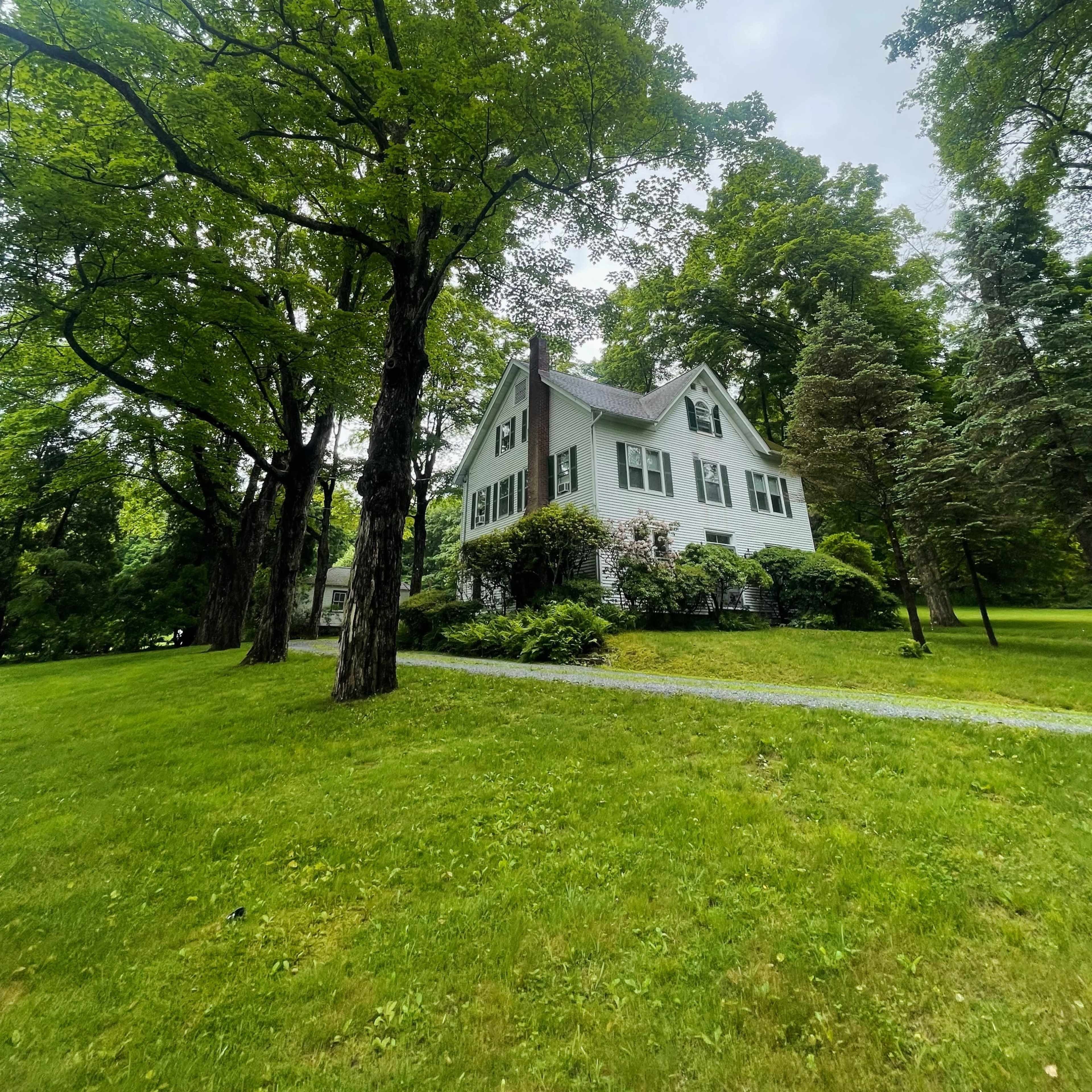 A white house with green shutters sits among tall trees and a well-maintained lawn.