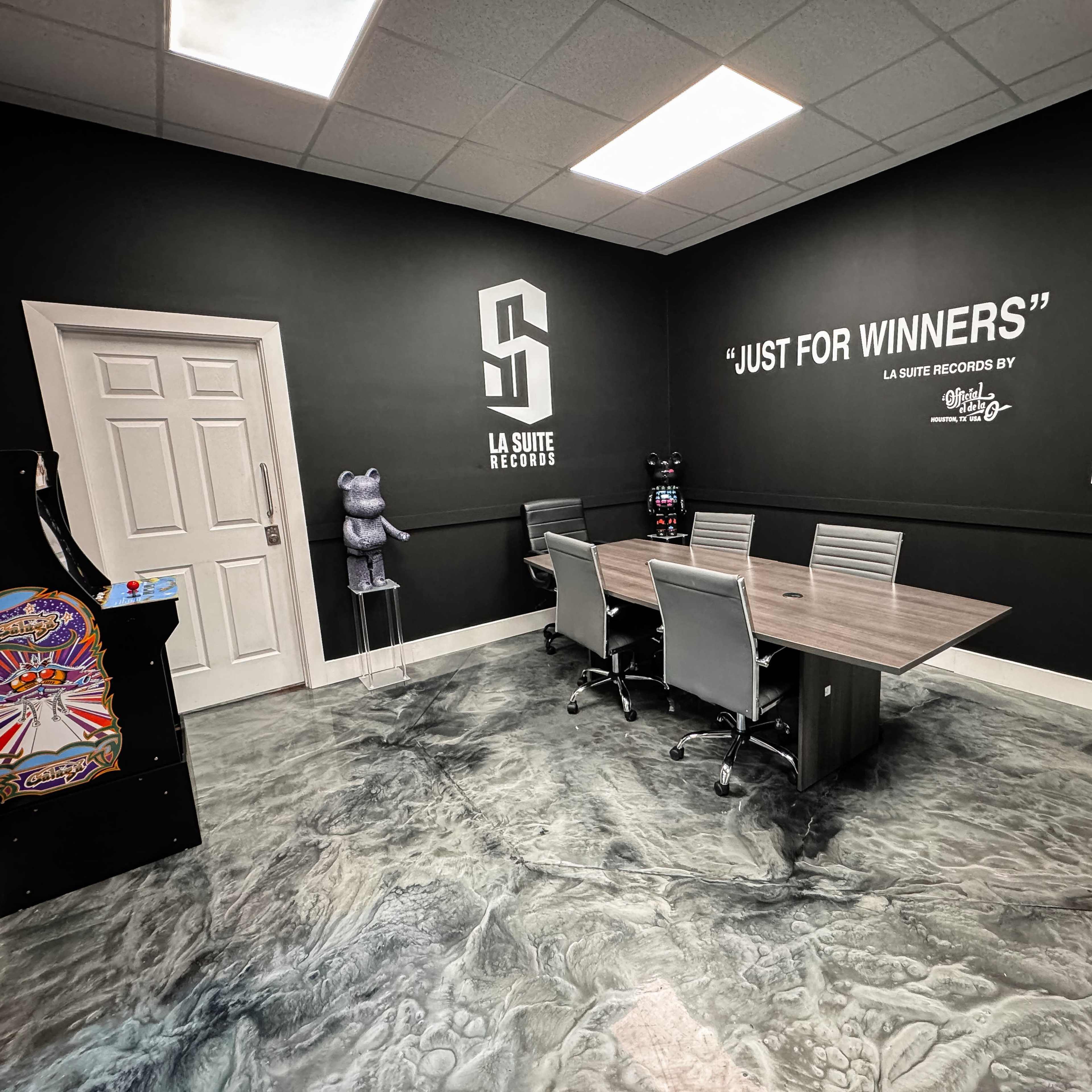 A conference room features a large table surrounded by chairs, with a black wall displaying the phrase "JUST FOR WINNERS" and an arcade machine in the corner.