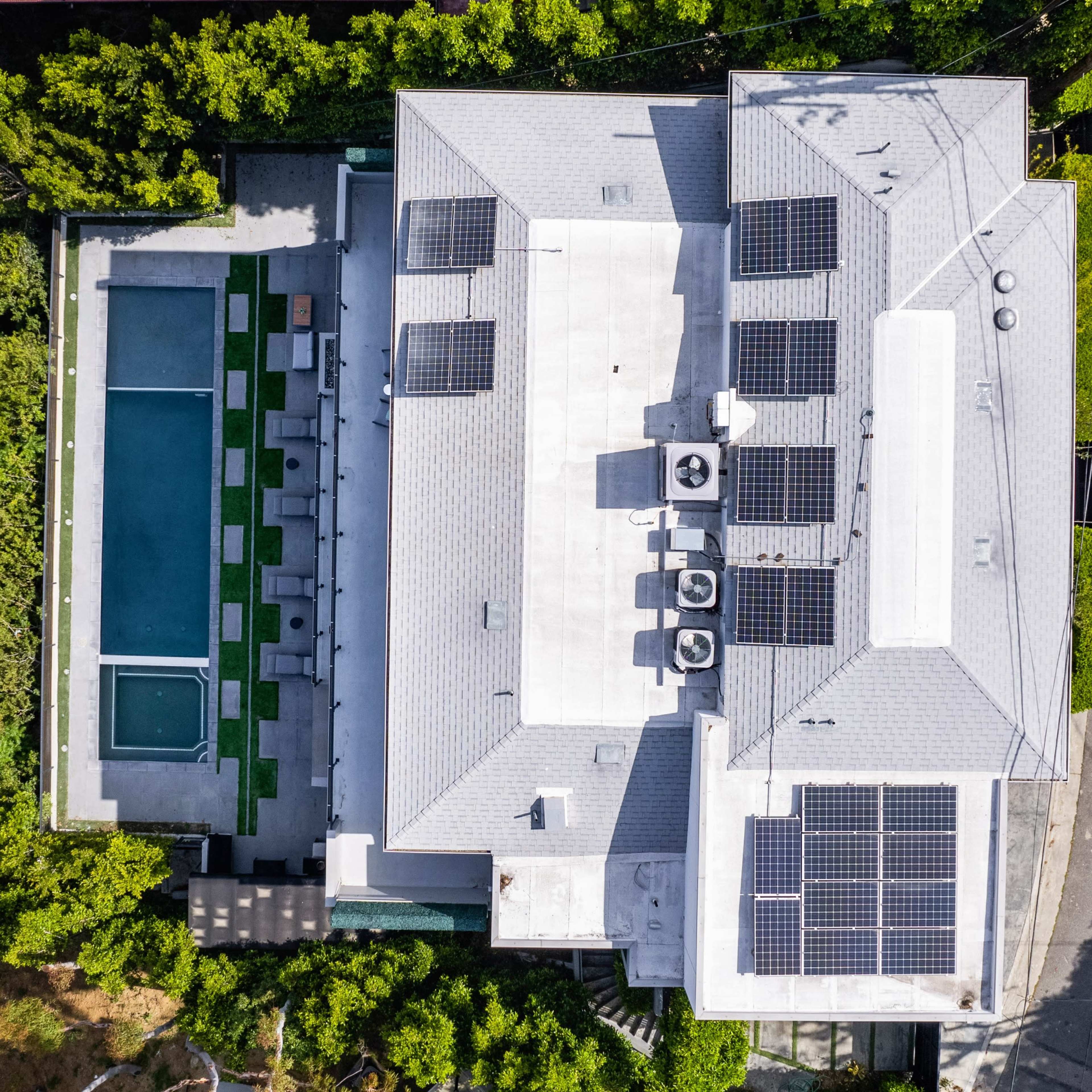 An aerial view of a modern house featuring solar panels on the roof, a swimming pool in the backyard, and surrounded by greenery.