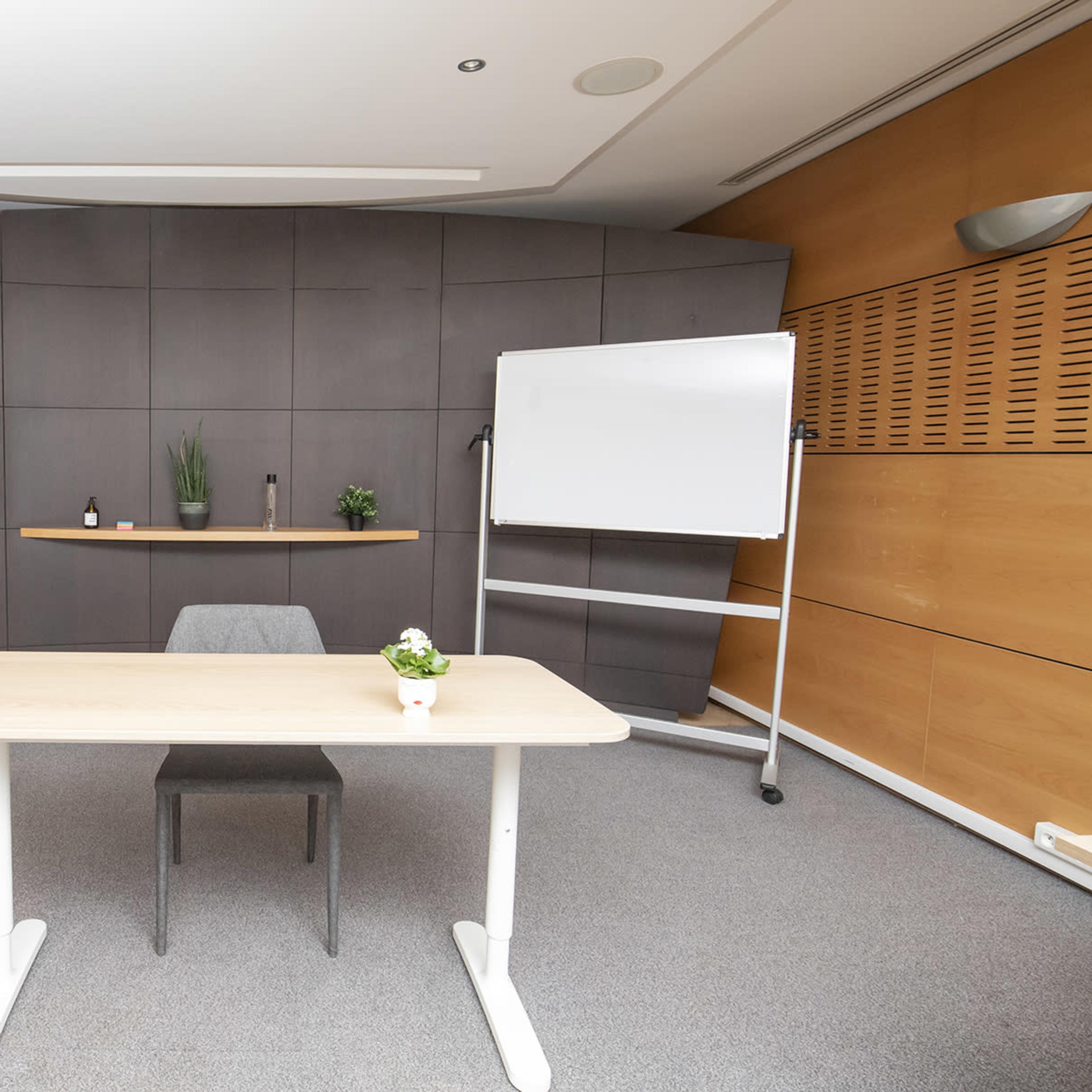 A minimalist meeting room featuring a table, a chair, a TV, a whiteboard, and a plant on a shelf against a paneled wall.