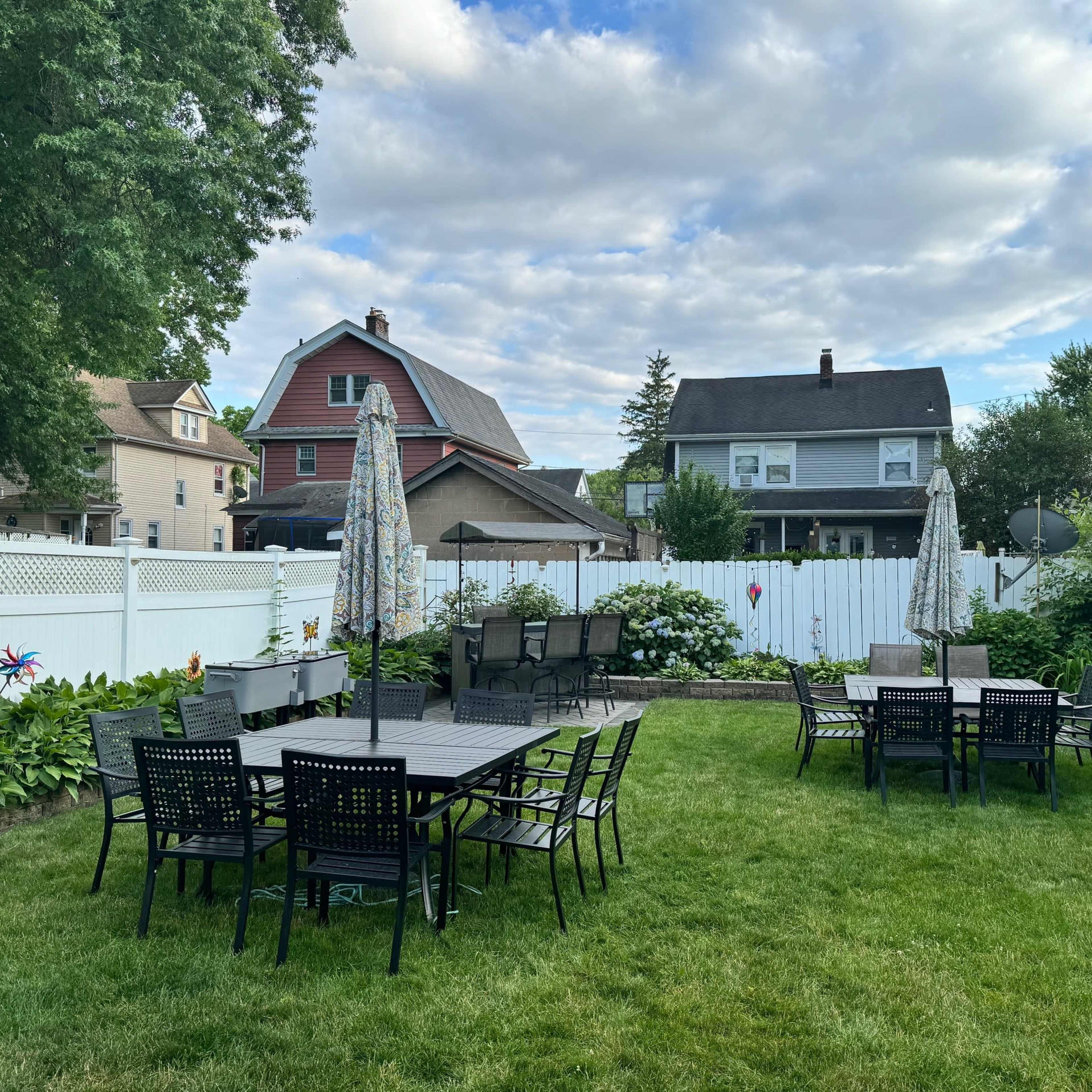 A backyard with several black patio tables and chairs arranged on a green lawn, surrounded by a white fence and various houses in the background.
