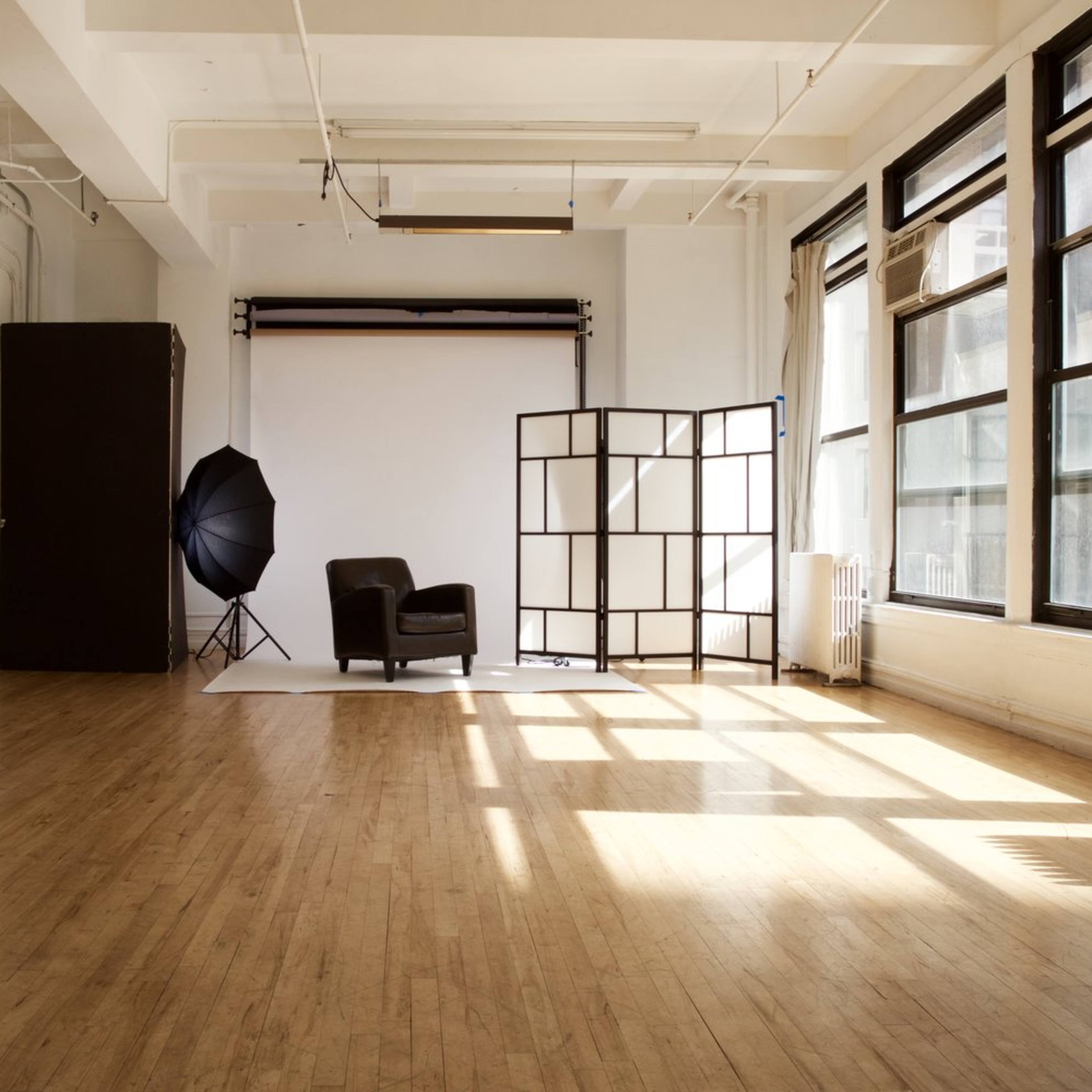 A bright, open studio space features a black chair, a backdrop setup, and large windows casting shadows on the wooden floor.