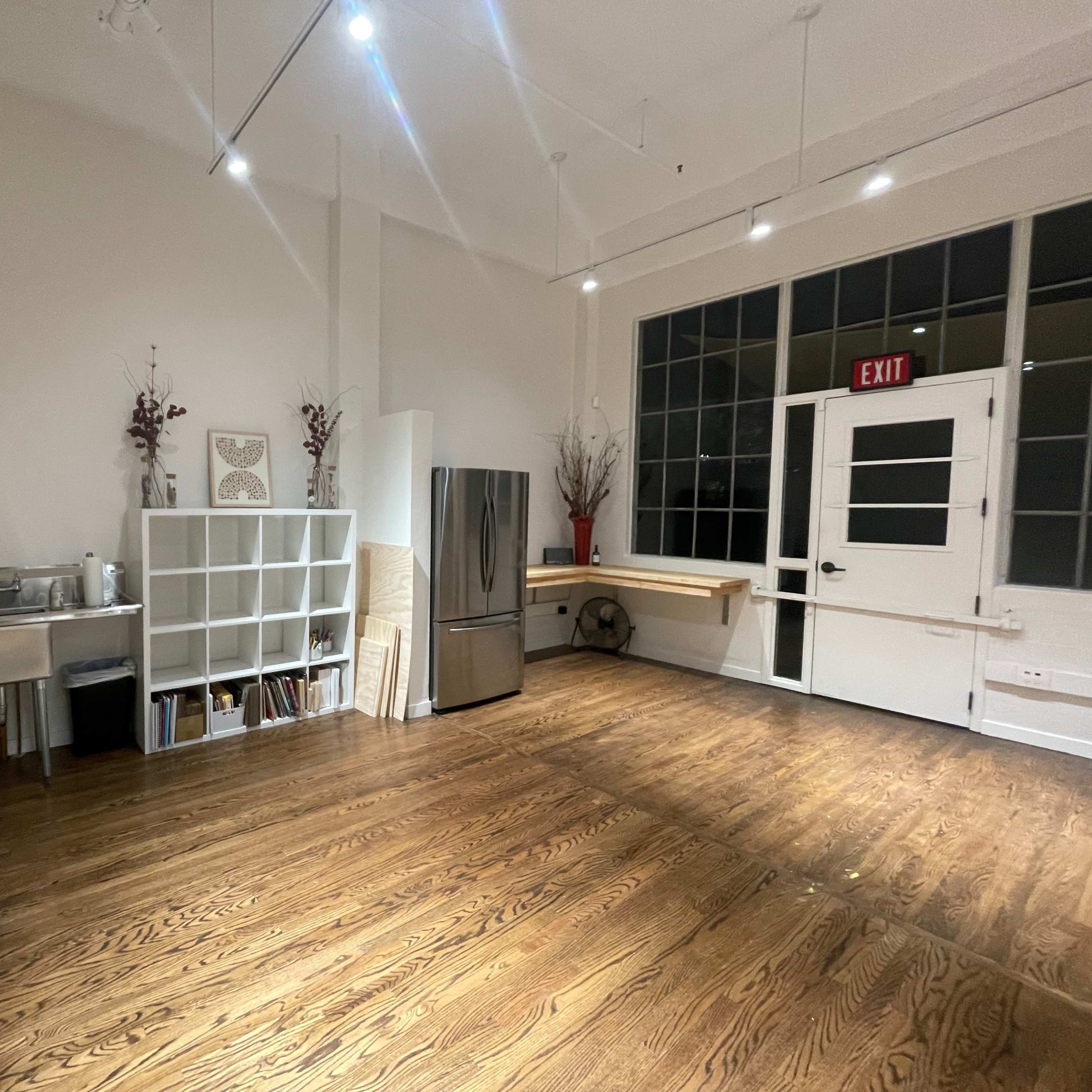 A spacious room with a wooden floor, a stainless steel refrigerator, a white shelving unit, and large windows.