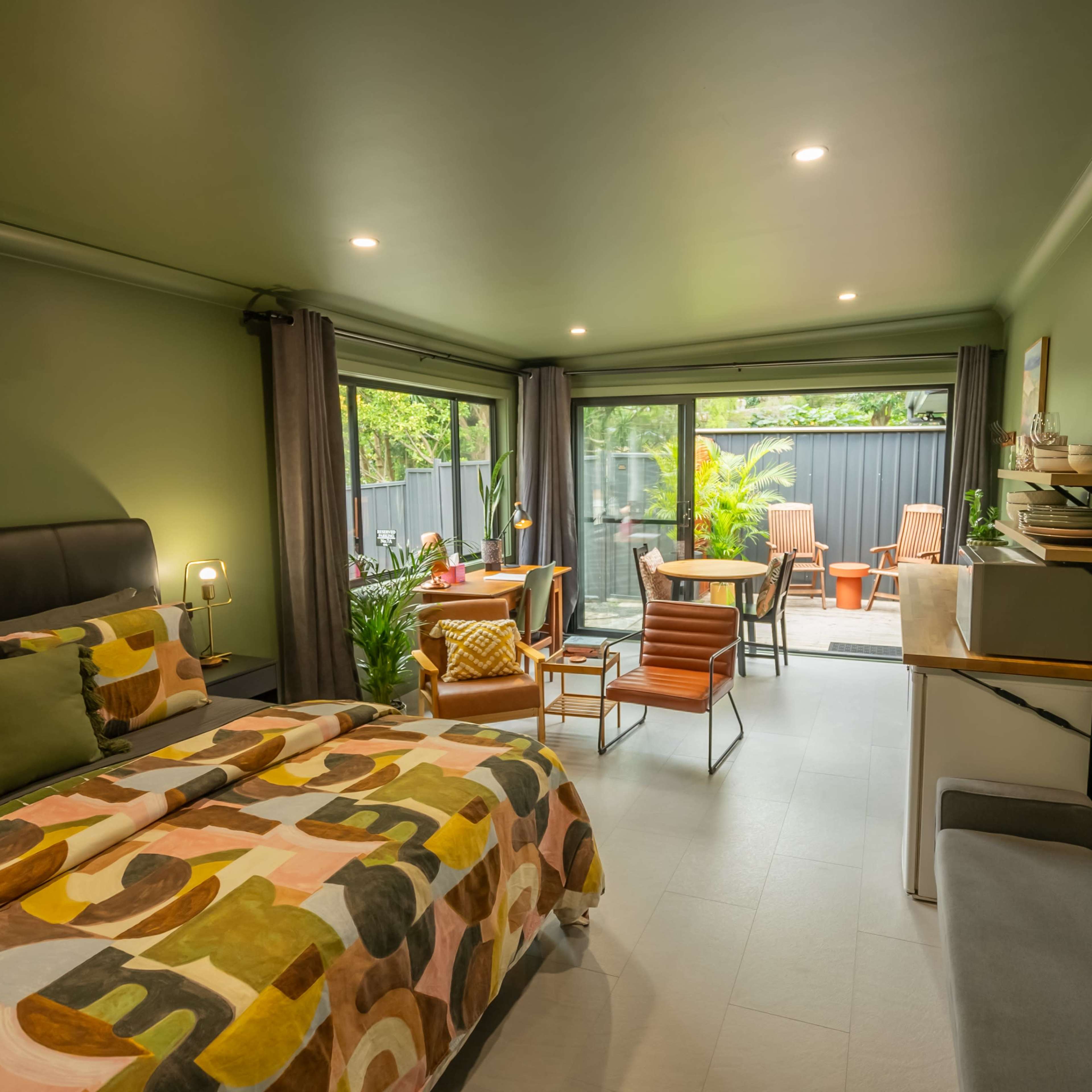 A modern bedroom features a bed with a colorful patterned duvet, seating areas, and large windows that open to a patio with outdoor furniture.