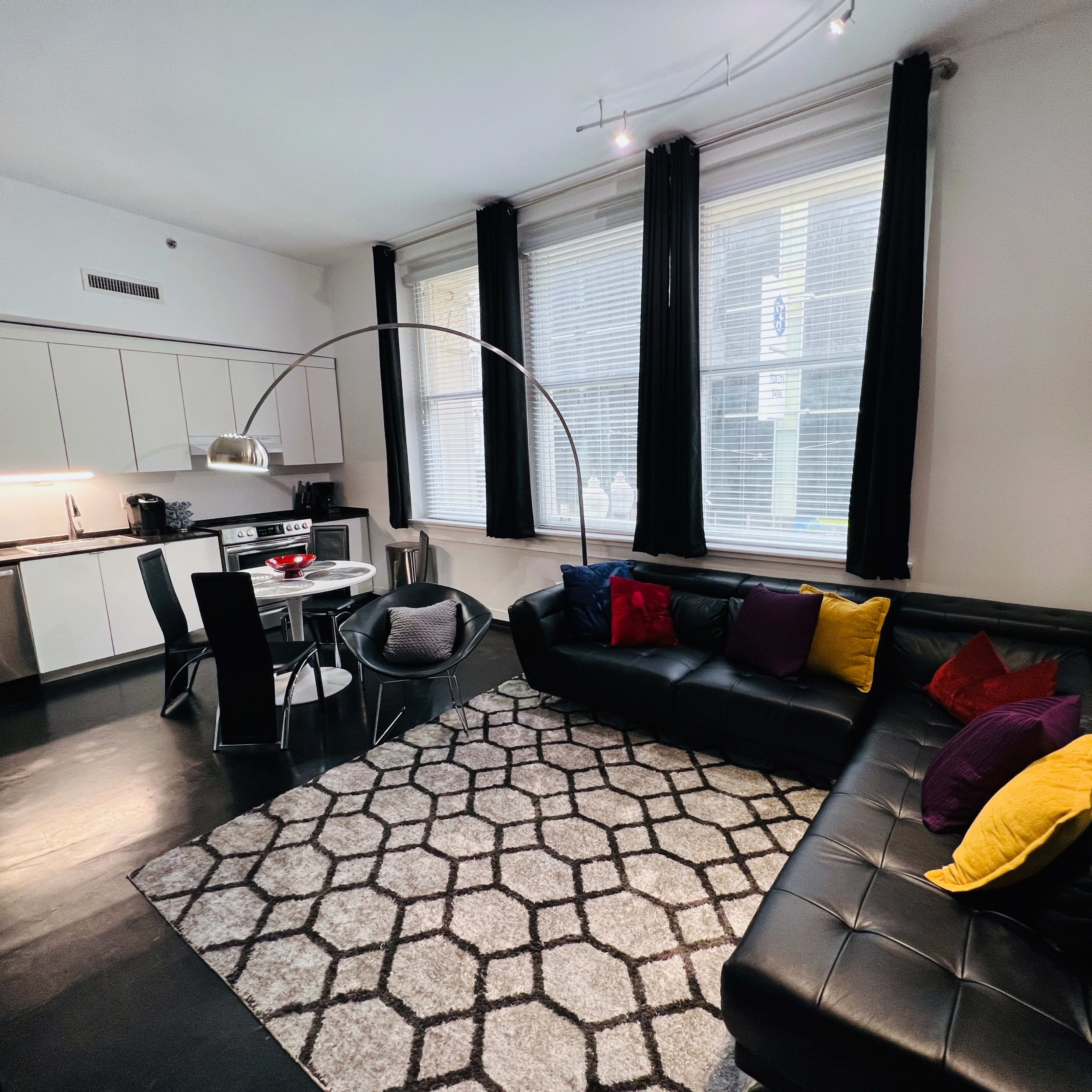A modern apartment living space with a black leather sectional, a round dining table, and large windows, adorned with a geometric rug and colorful pillows.