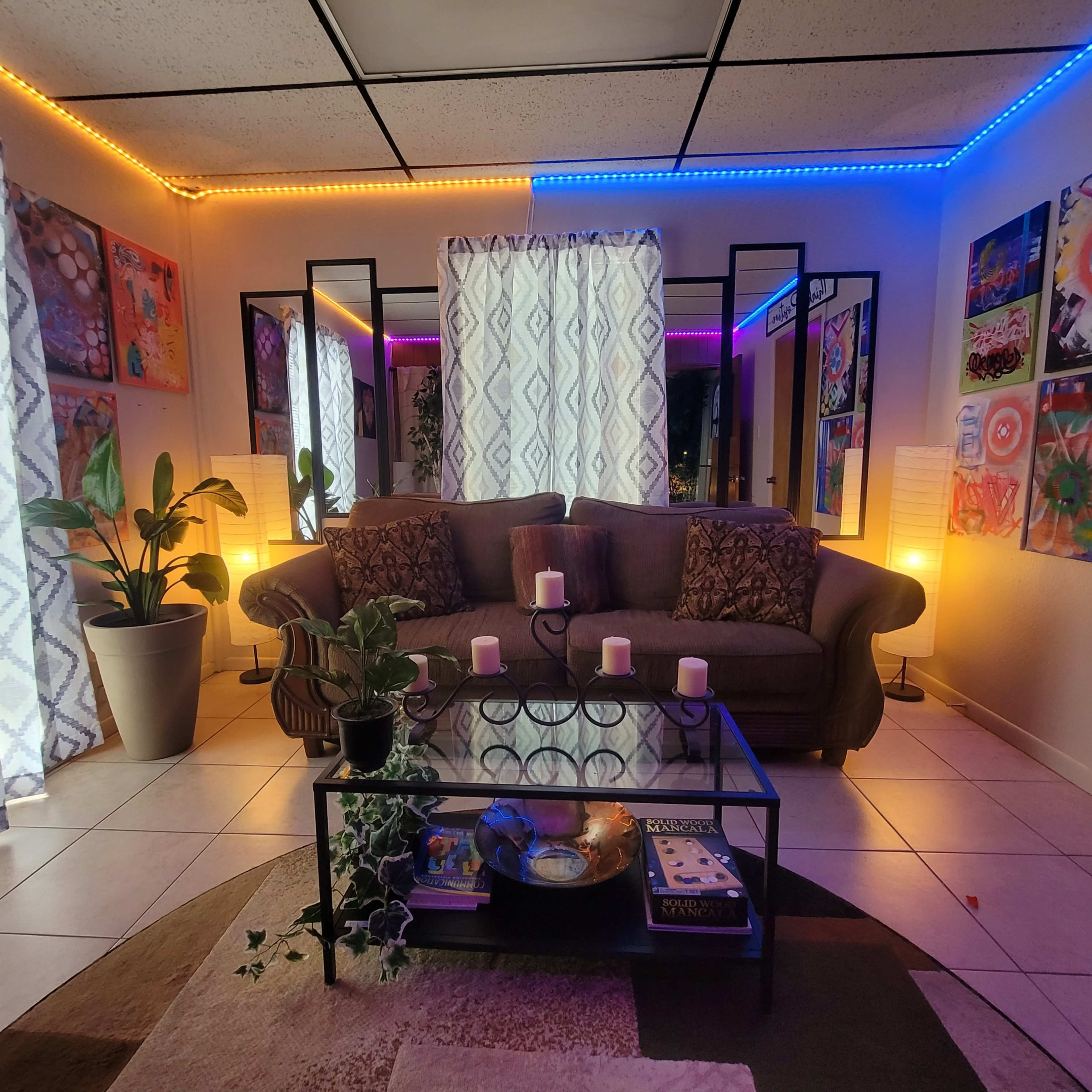 A living room with a gray sofa, a glass coffee table, and colorful LED lights illuminating the walls, surrounded by artwork and plants.
