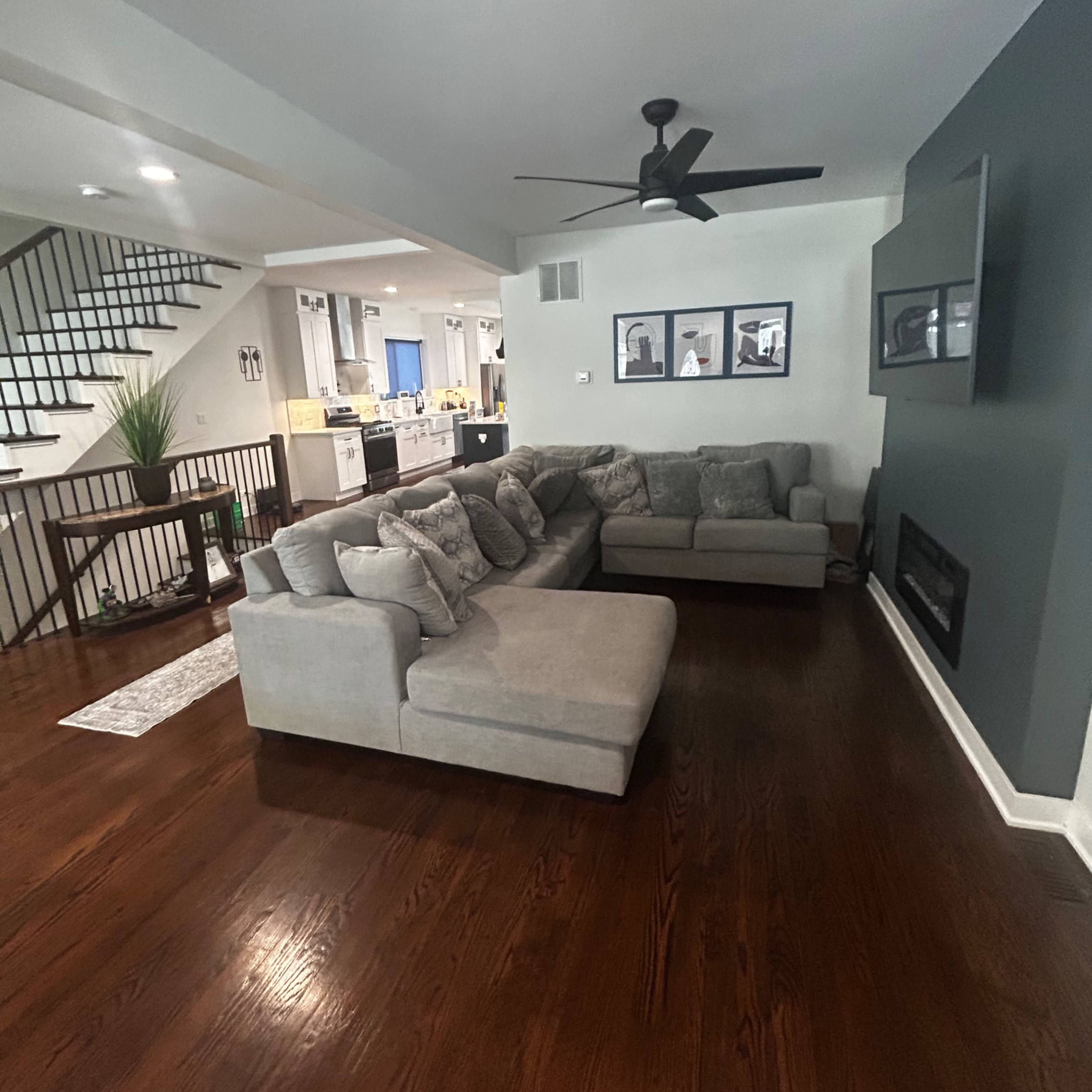 A modern living room featuring a large gray sectional sofa, a wall-mounted television, and a staircase in the background.
