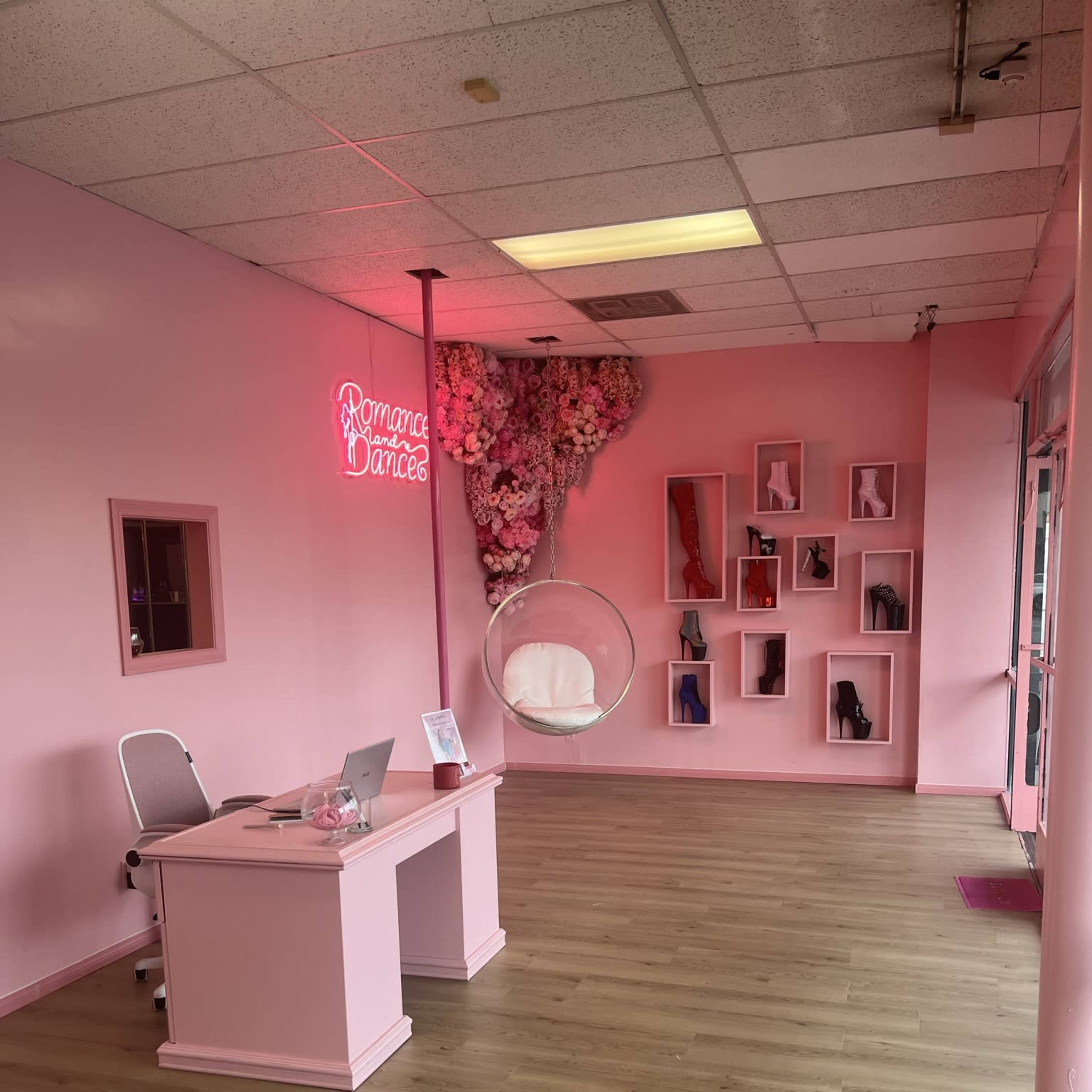 A pink-themed interior of a dance studio with a neon sign, a desk with a laptop, and a hanging bubble chair beside a wall adorned with high-heeled shoes.