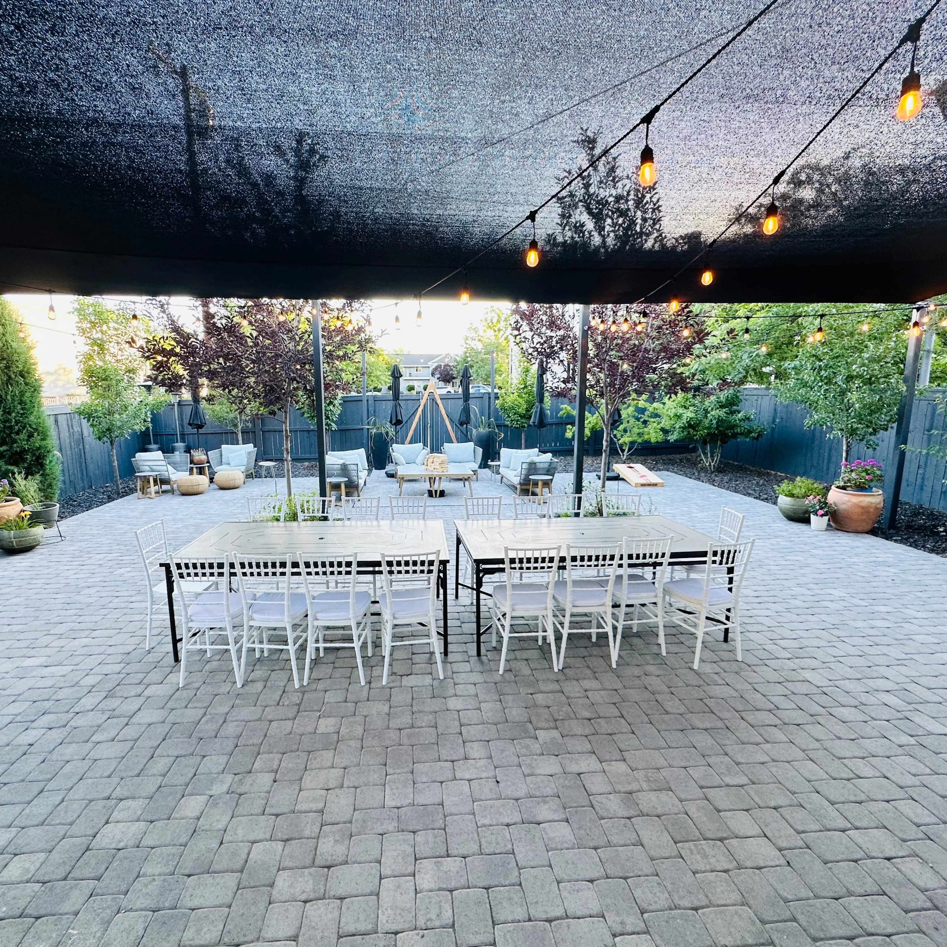 A spacious outdoor patio area with a long dining table and white chairs arranged under string lights and a shaded canopy.