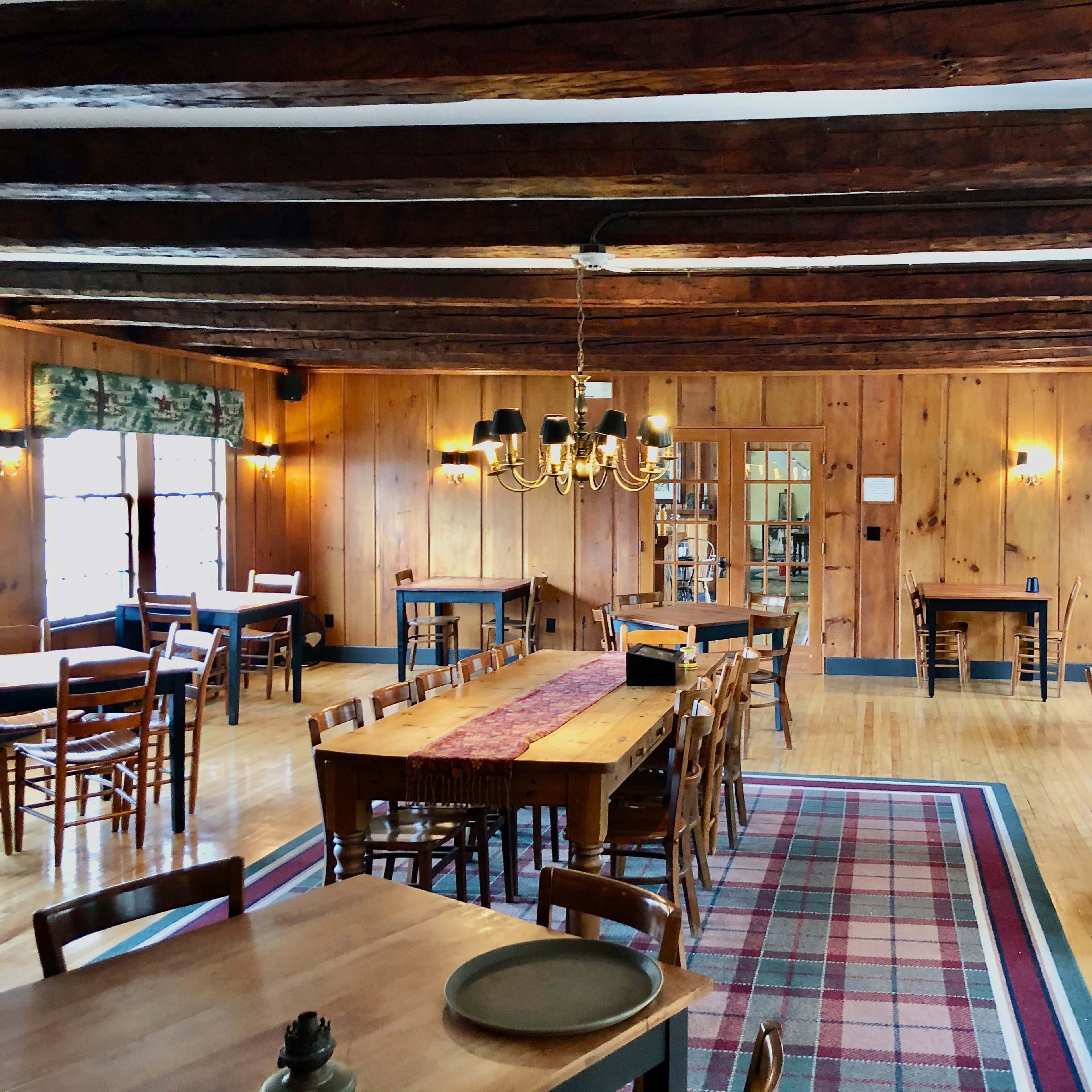 A wooden-paneled dining room with multiple tables, a chandelier, and large windows letting in natural light.