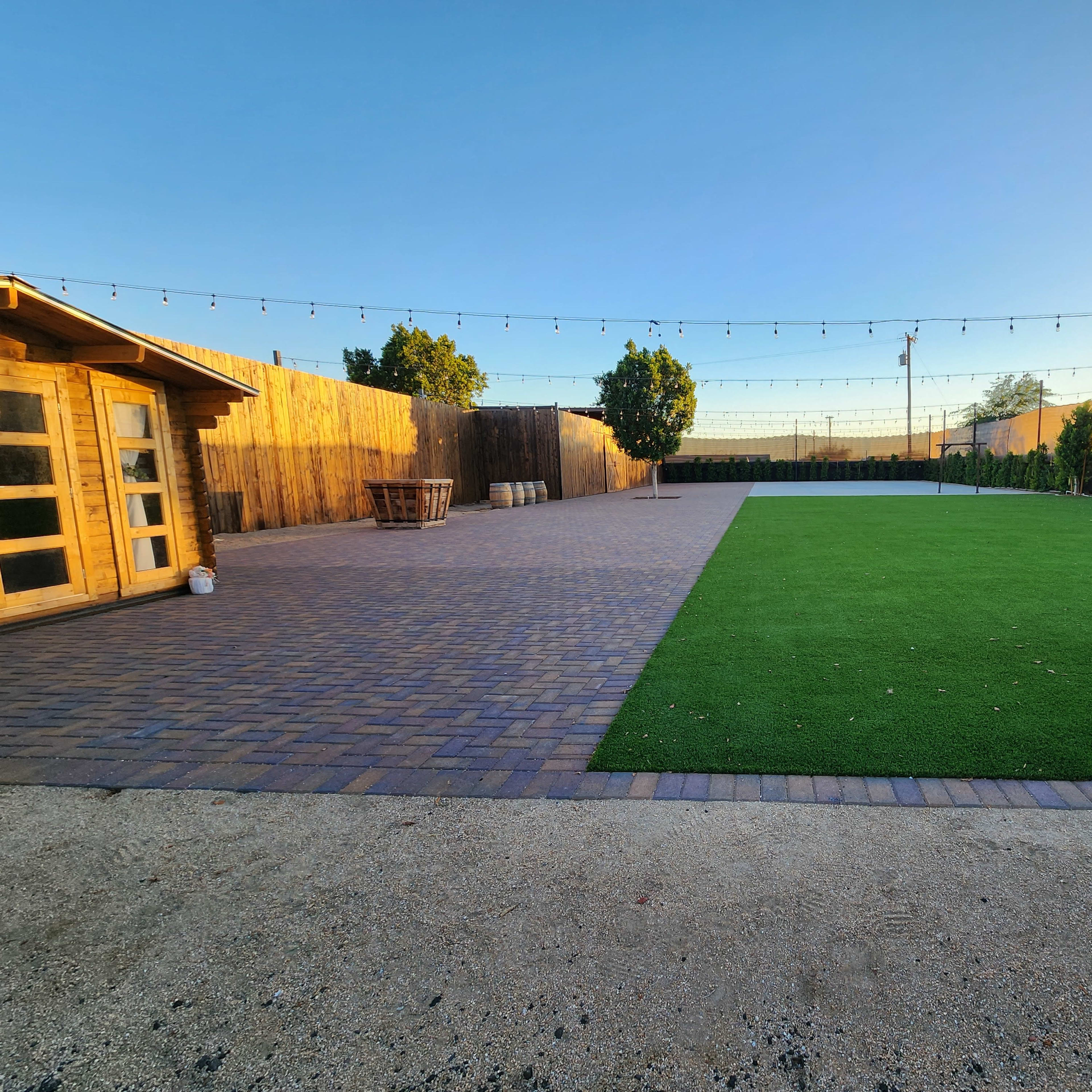A spacious backyard with a section of artificial grass, a patio made of pavers, and wooden fencing along the perimeter, illuminated by string lights overhead.