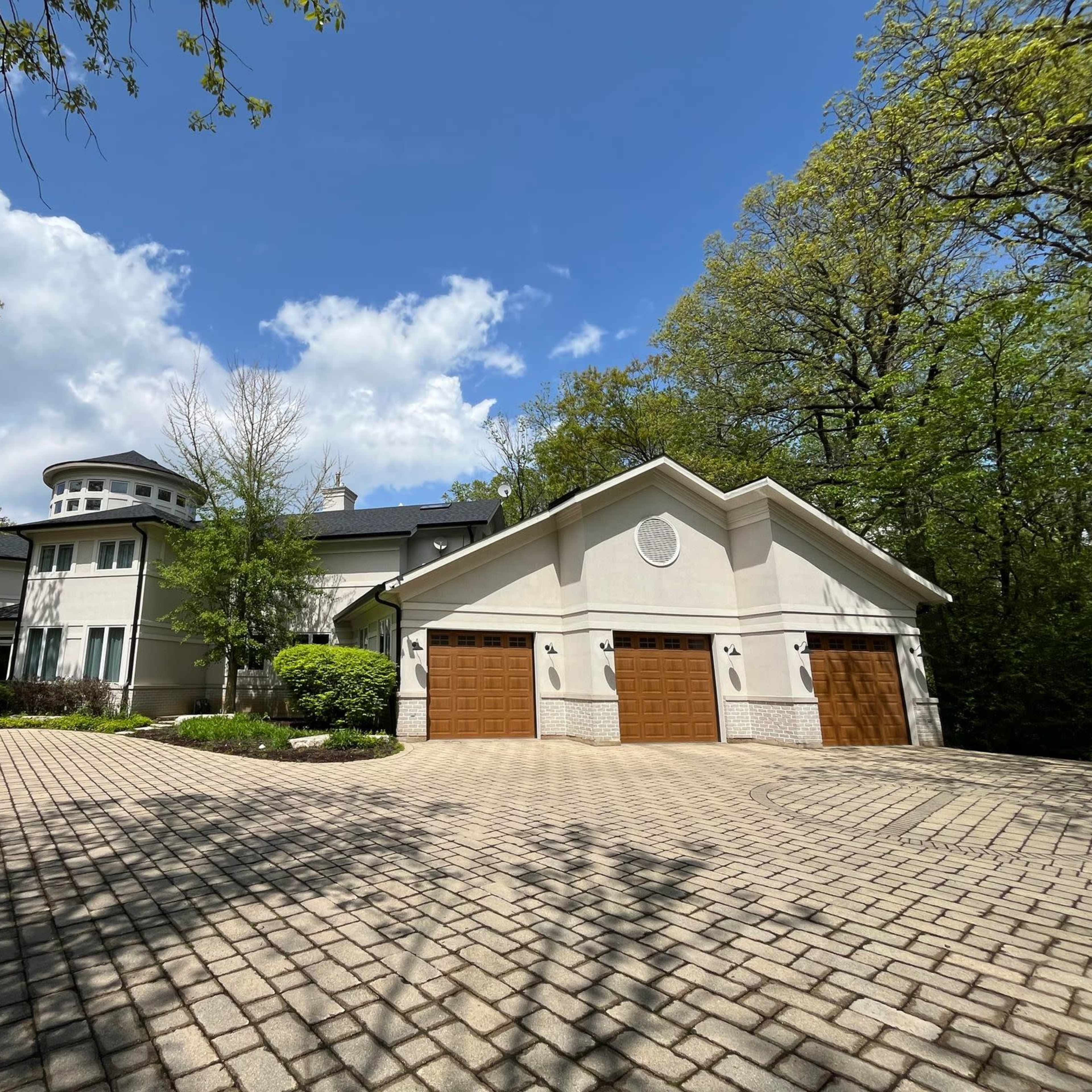A large, modern house with a circular turret and a three-car garage is set among trees under a partly cloudy sky.