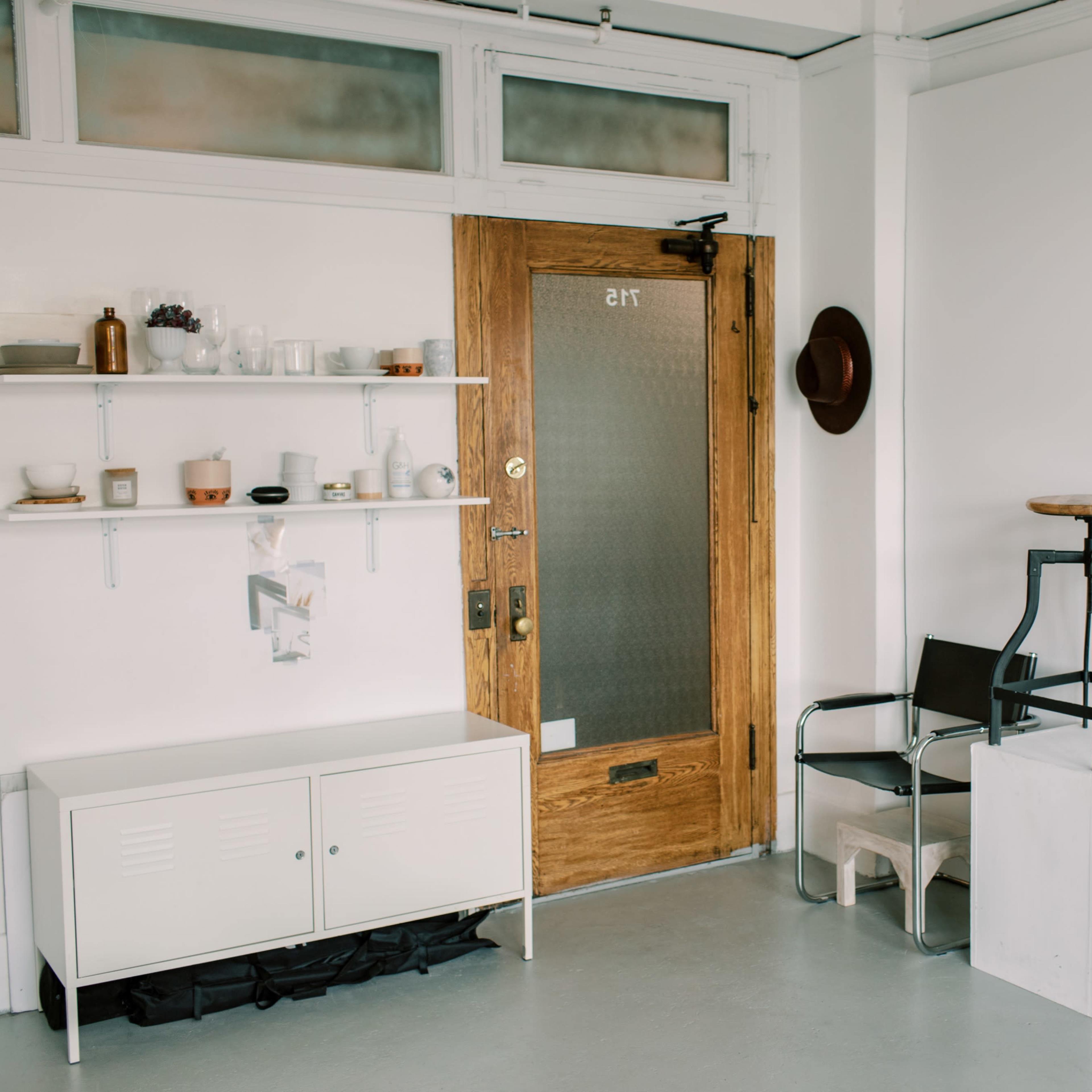 A minimalist room with a wooden door, a wall-mounted shelf filled with items, a black chair, a white stool, and a full-length mirror.
