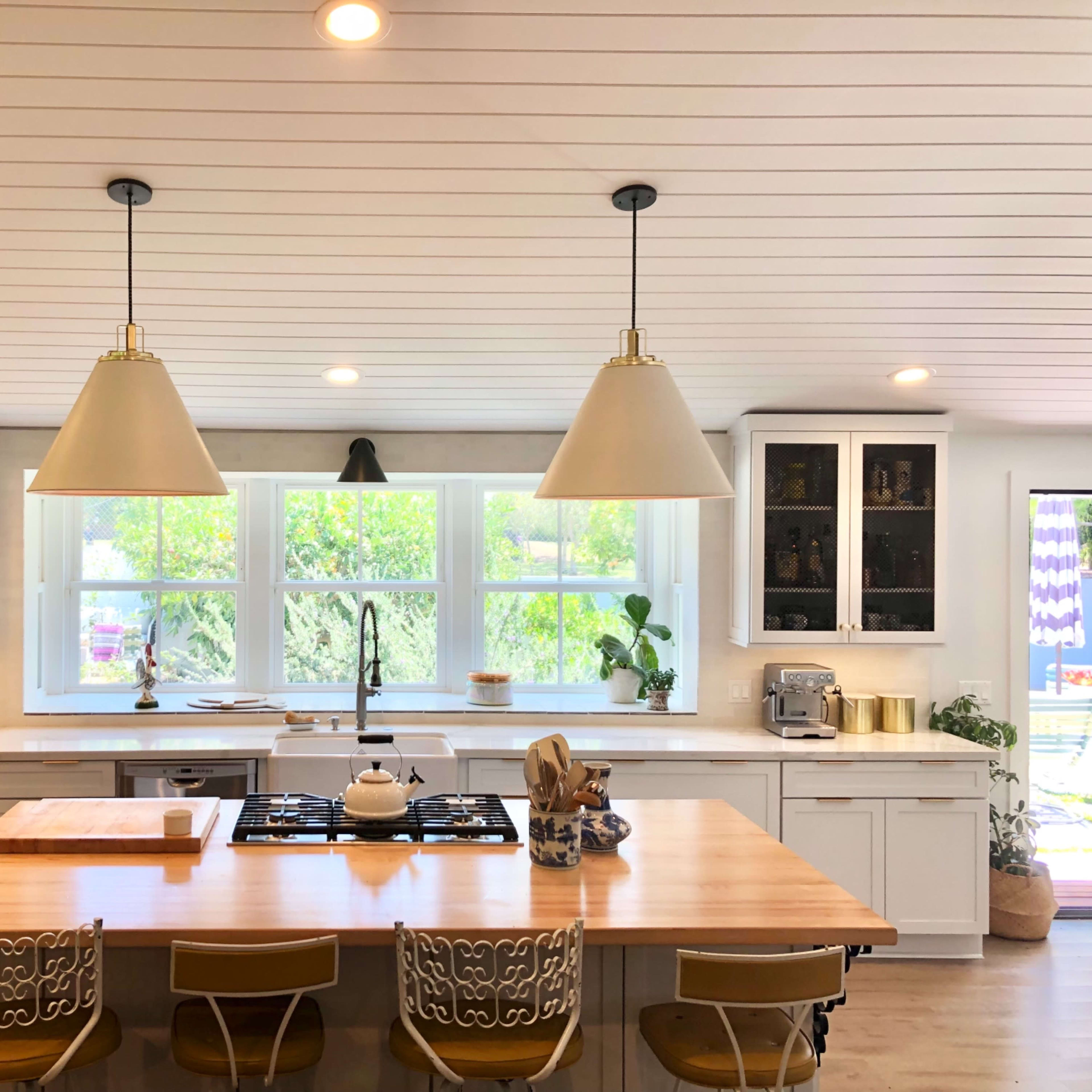 A modern kitchen featuring a central island with a cooktop, pendant lights, and white cabinetry, illuminated by natural light from large windows.