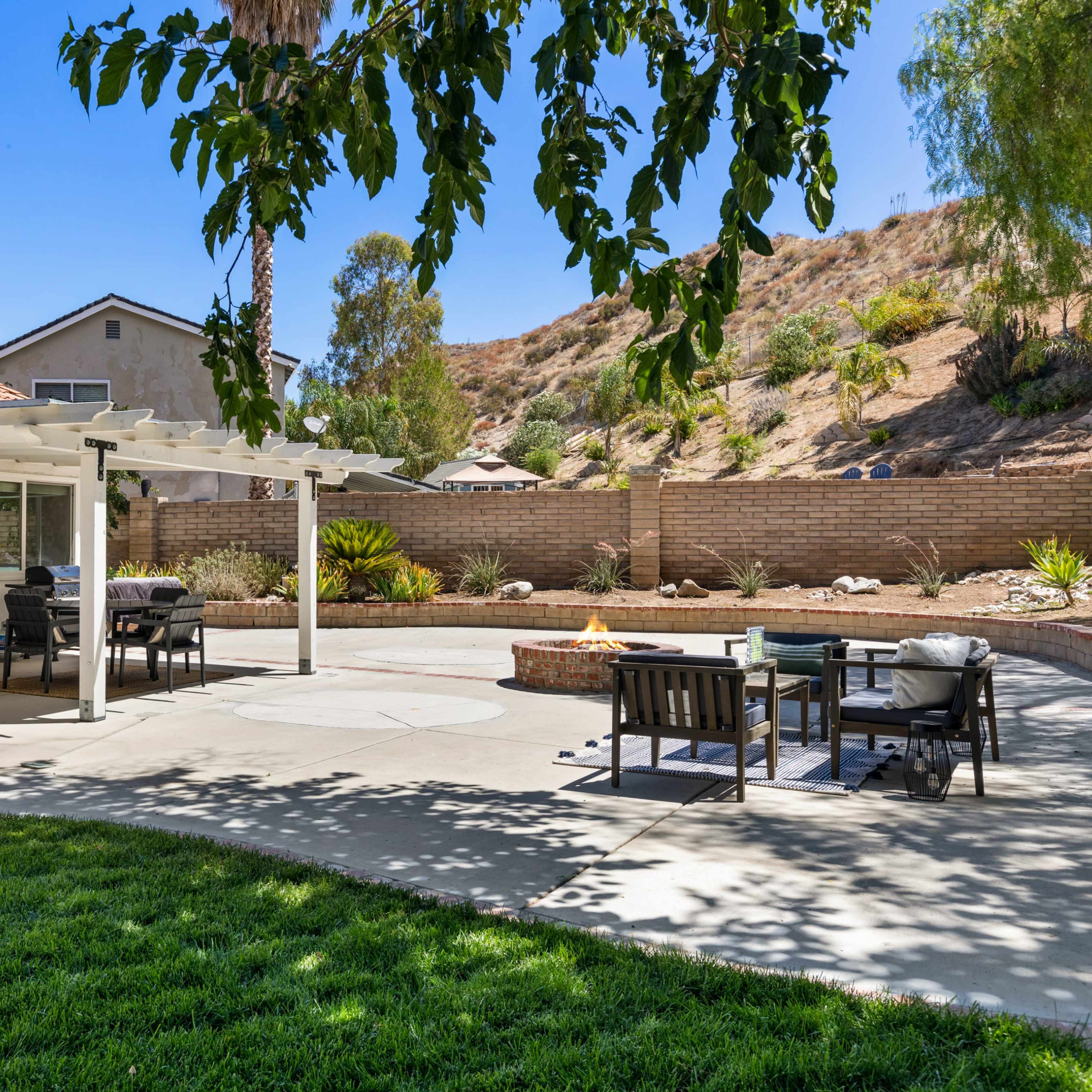 A patio area features a pergola, outdoor seating, and a fire pit, surrounded by a lush green lawn and hillside.