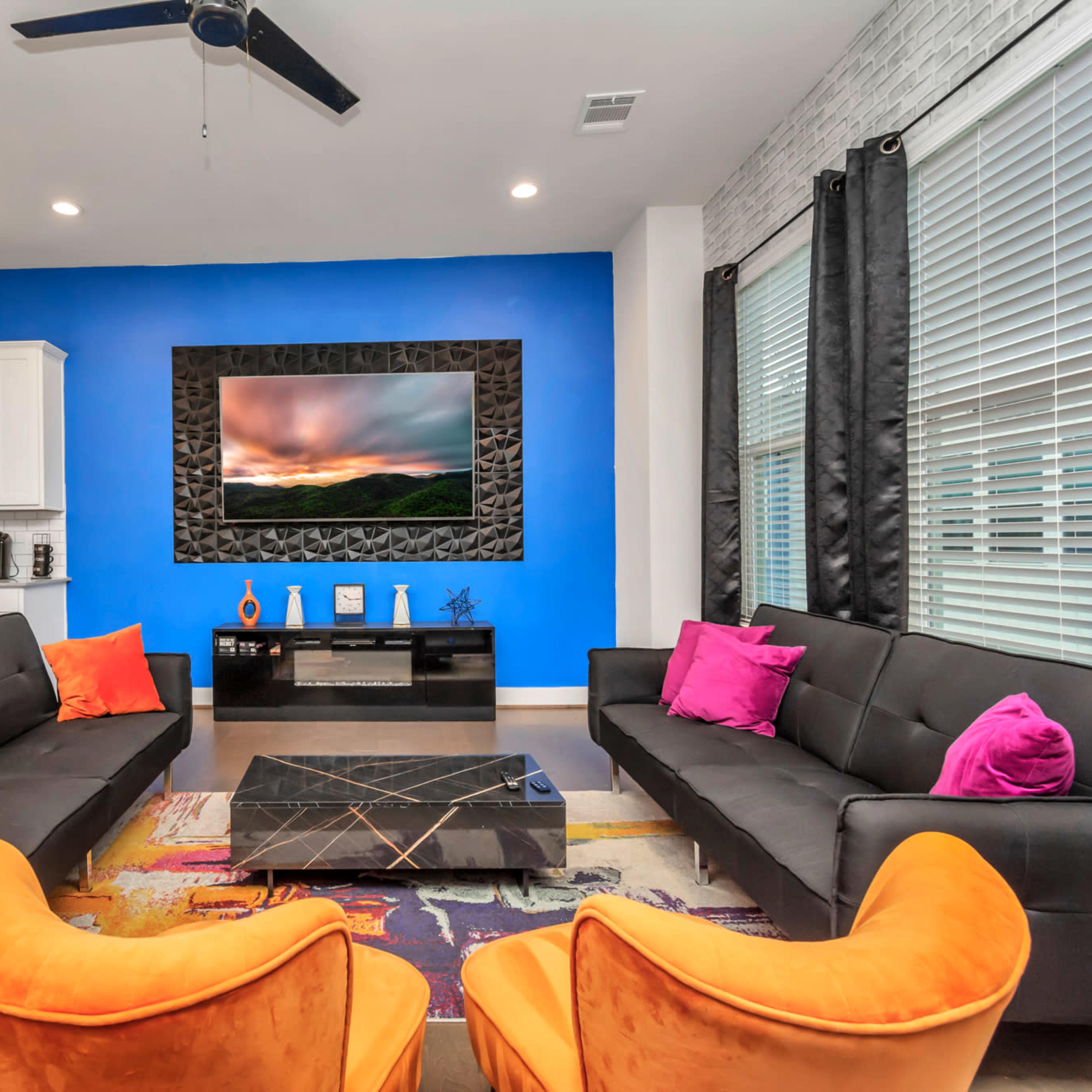 A modern living room features a blue accent wall, two black sofas with colorful cushions, an orange armchair, a coffee table, and a mounted television above a sleek fireplace.