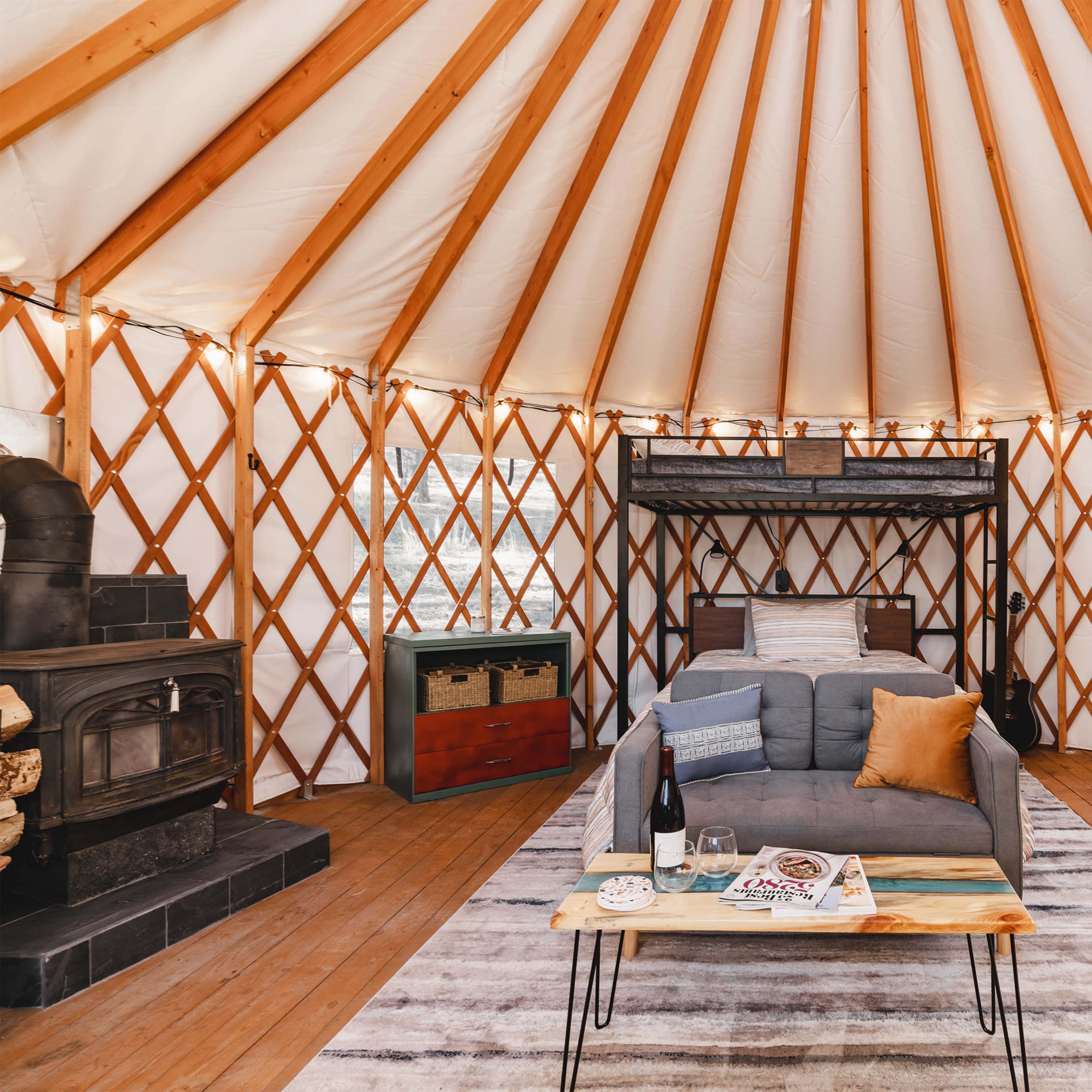 The interior of a yurt with wooden beams, a sofa, a bed loft, and a coffee table arranged on a striped rug.