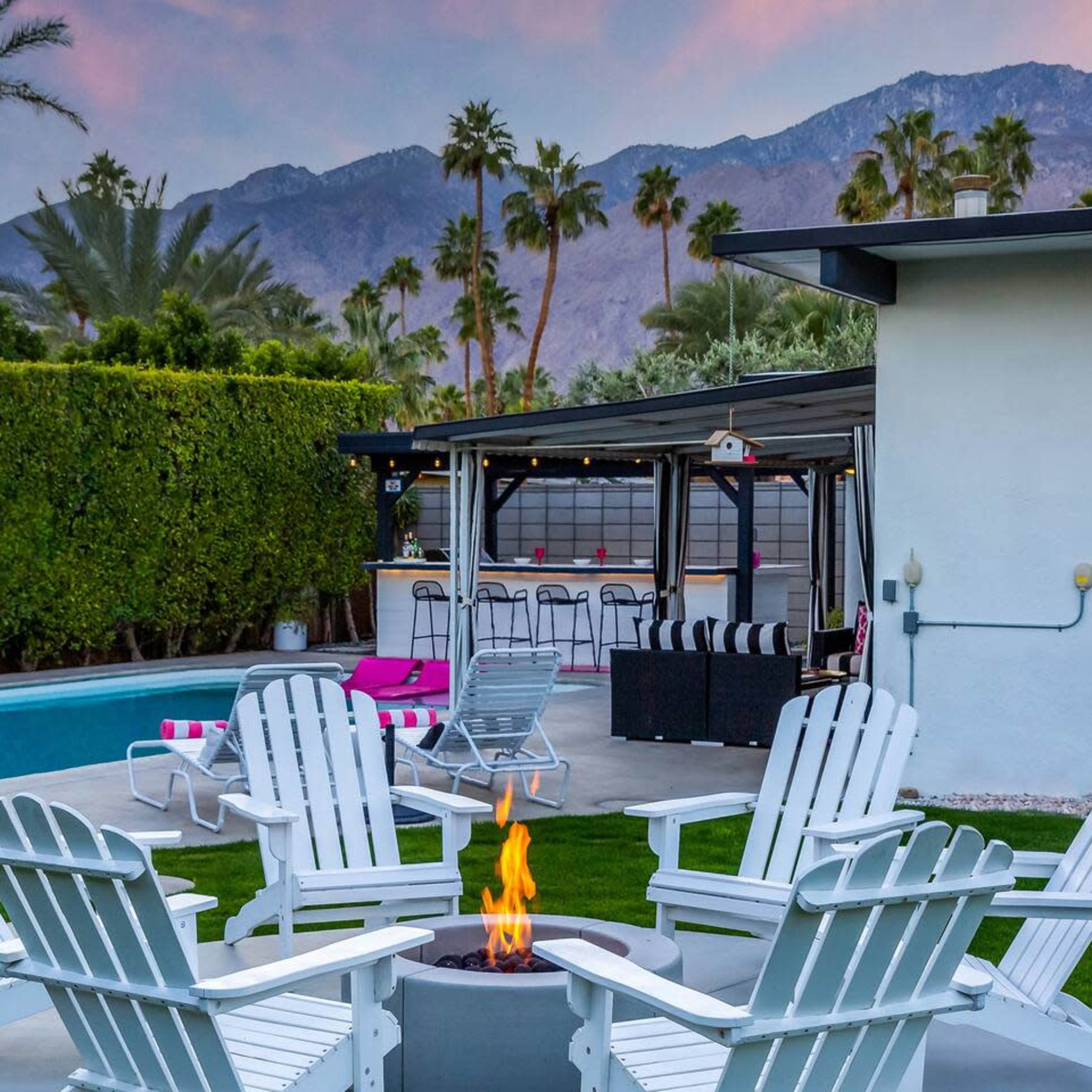 A circular arrangement of white chairs surrounds a fire pit near a pool and a bar area, with mountains in the background and palm trees lining the property.