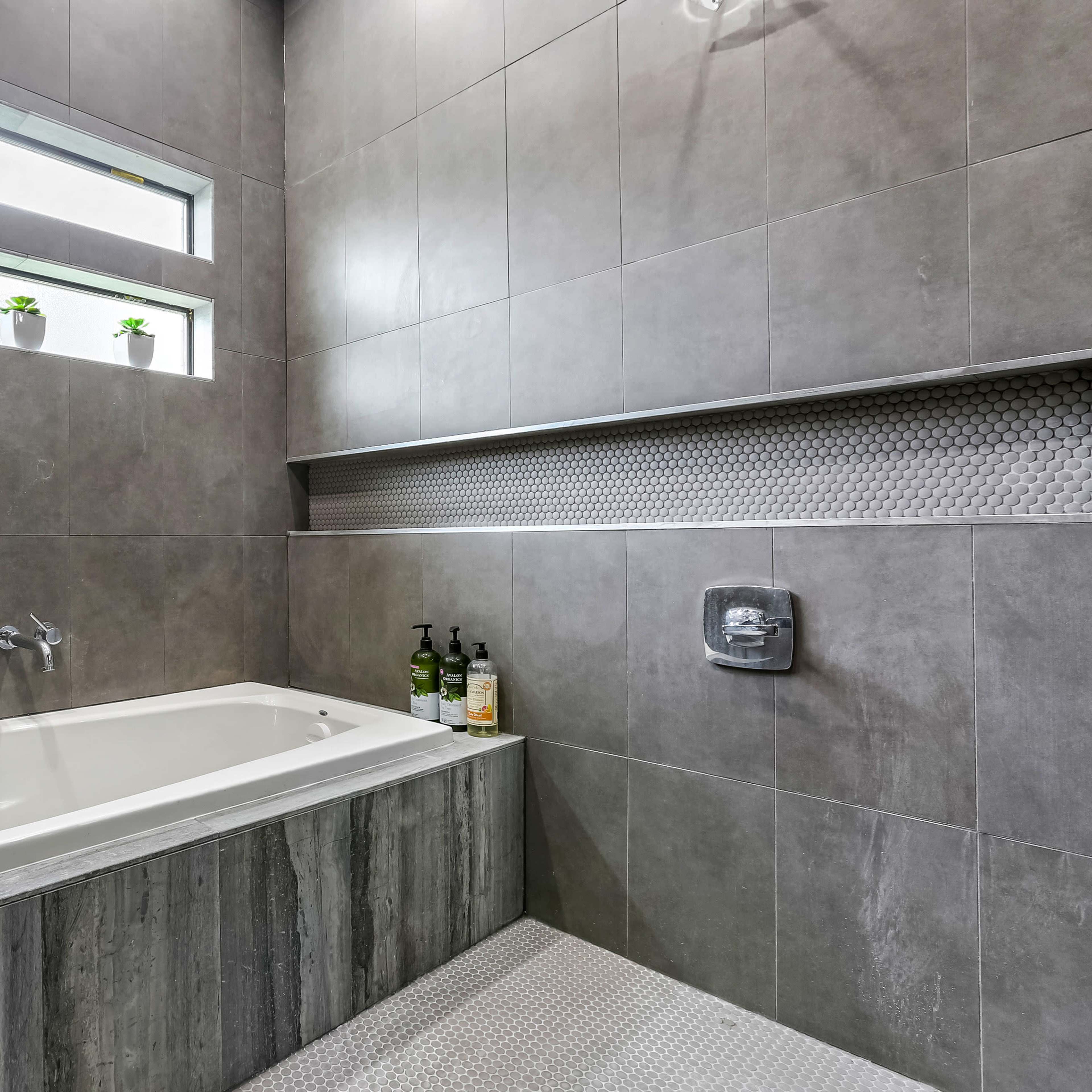 A modern bathroom with a freestanding bathtub, gray tiled walls, a narrow window, and a shelf holding several bottles.