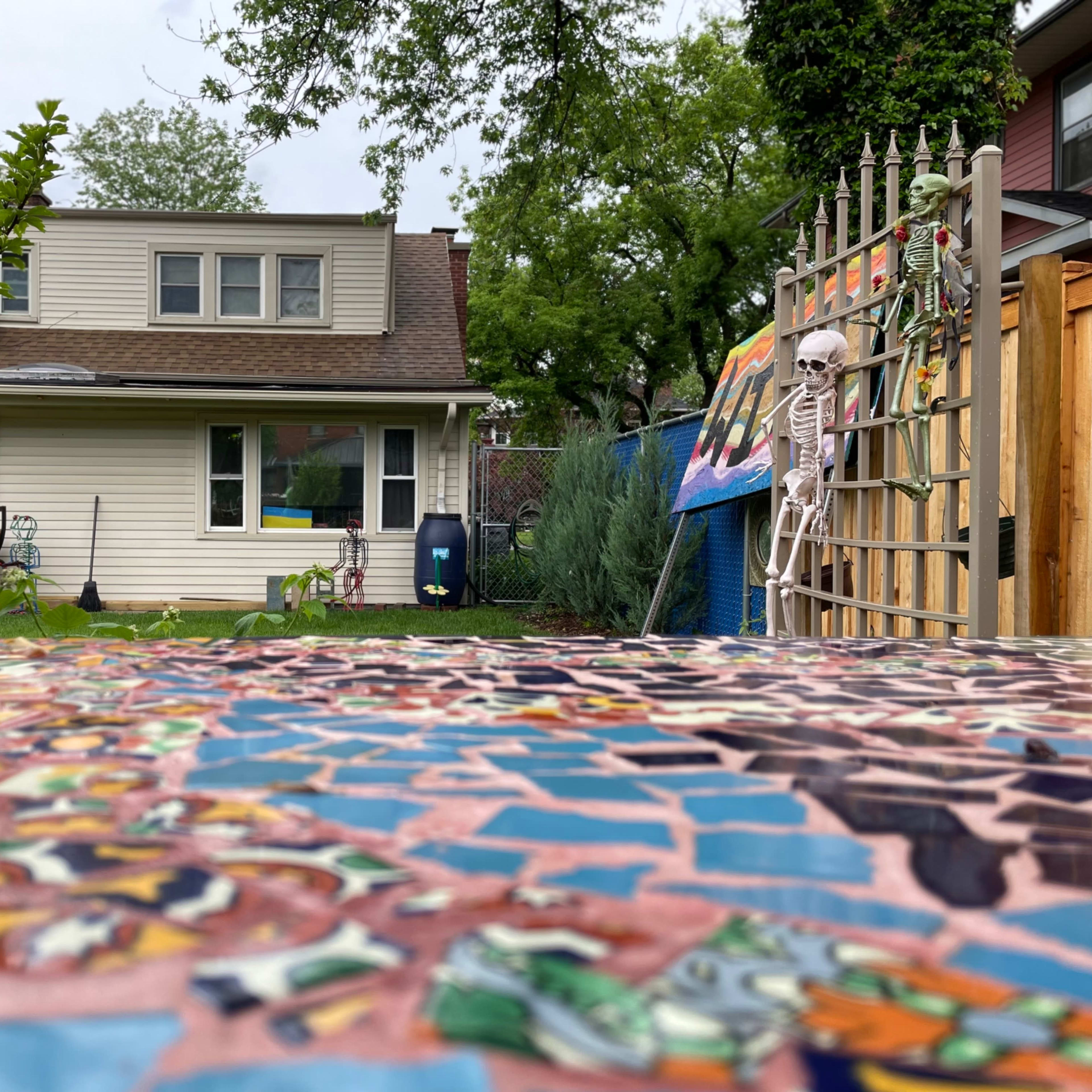 A backyard with a mosaic table in the foreground, a house in the background, and a skeleton decoration near a gate.