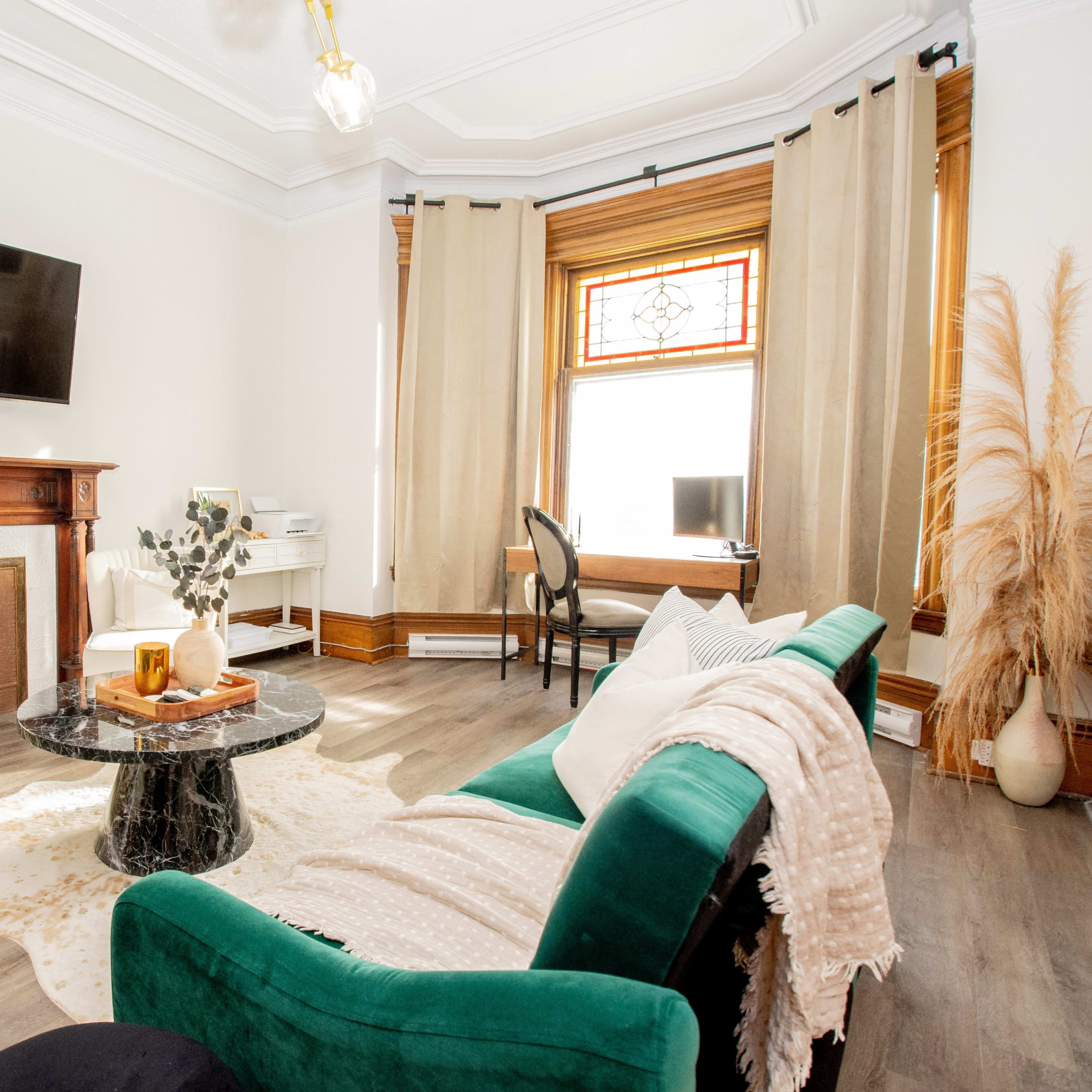The room features a cozy seating area with a green sofa, a coffee table, and a fireplace, along with a workspace in the background.