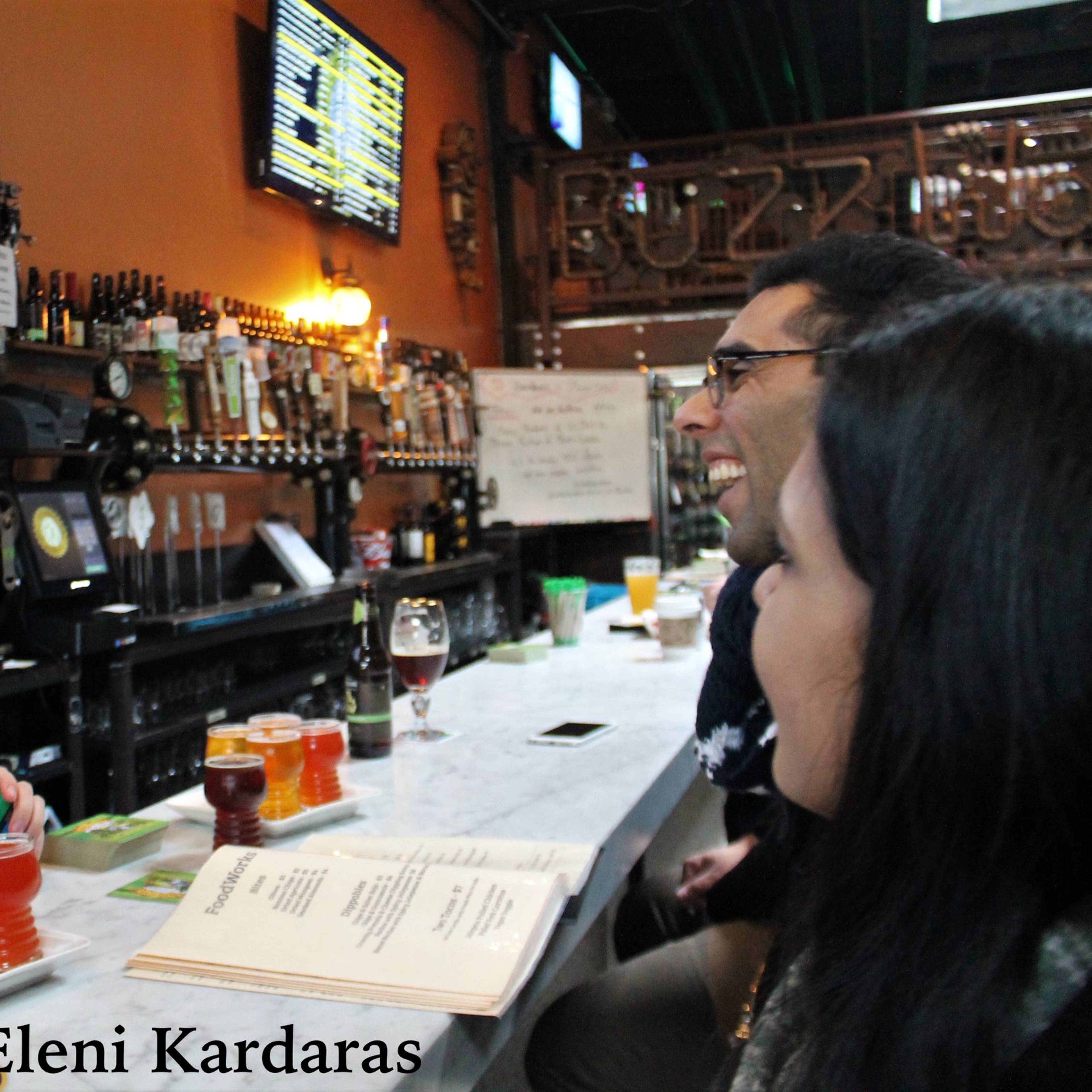 A bartender serves a variety of colorful beers to two customers at a bar with numerous drink taps in the background.