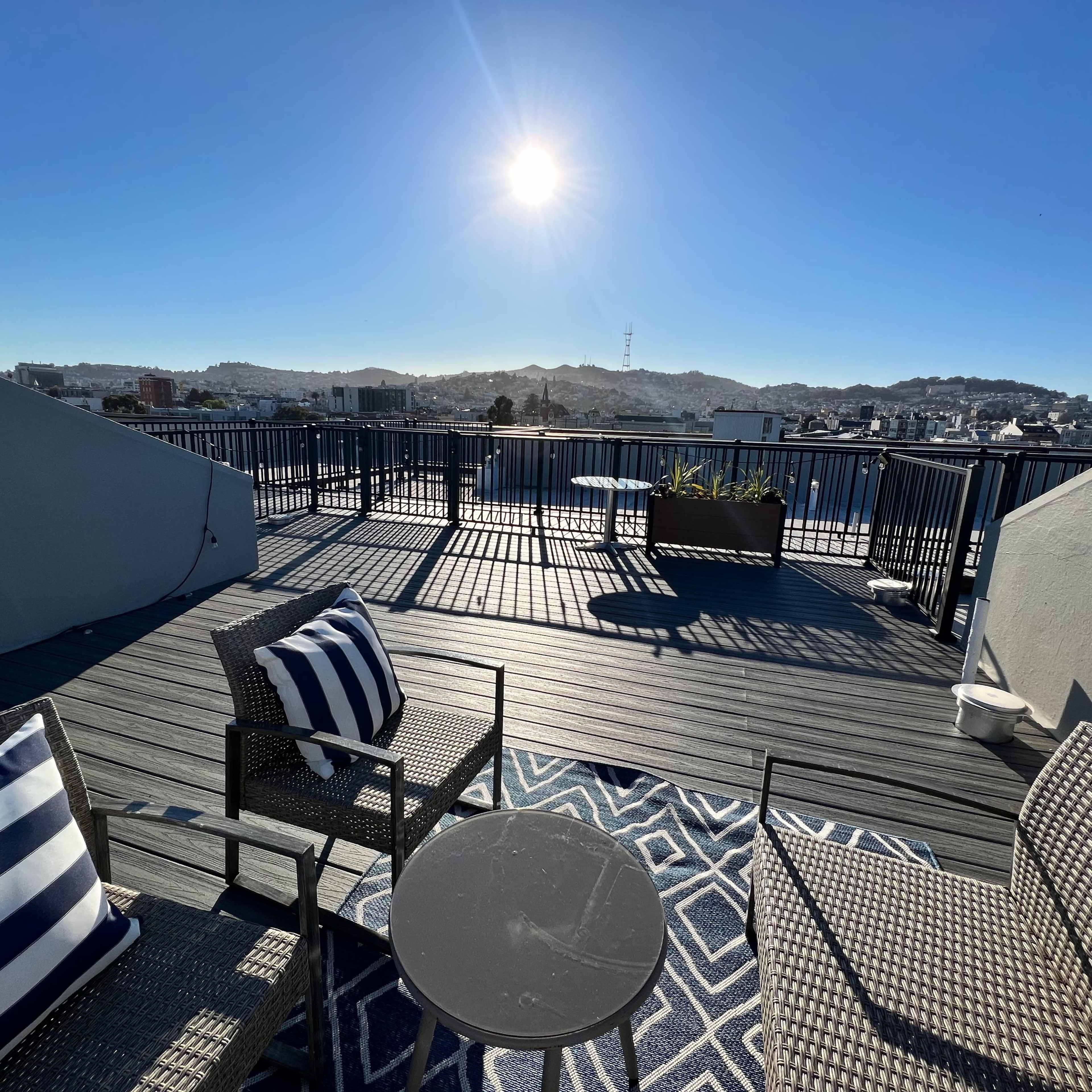 A rooftop terrace features two chairs with striped cushions, a small table, and a sun shining brightly over a cityscape.