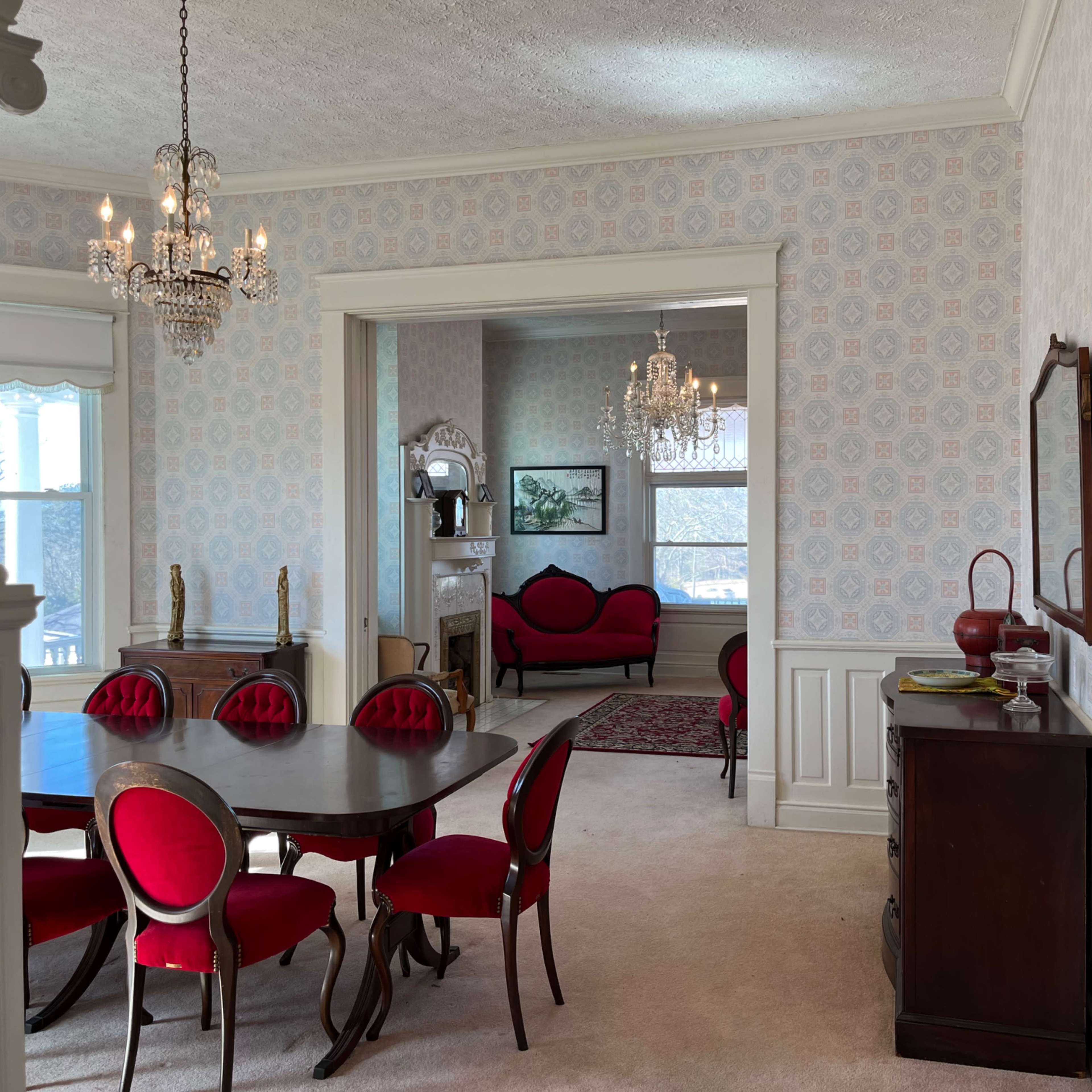 An elegantly furnished room with a dining table and red chairs, leading into a connected sitting area with couches and chandeliers.