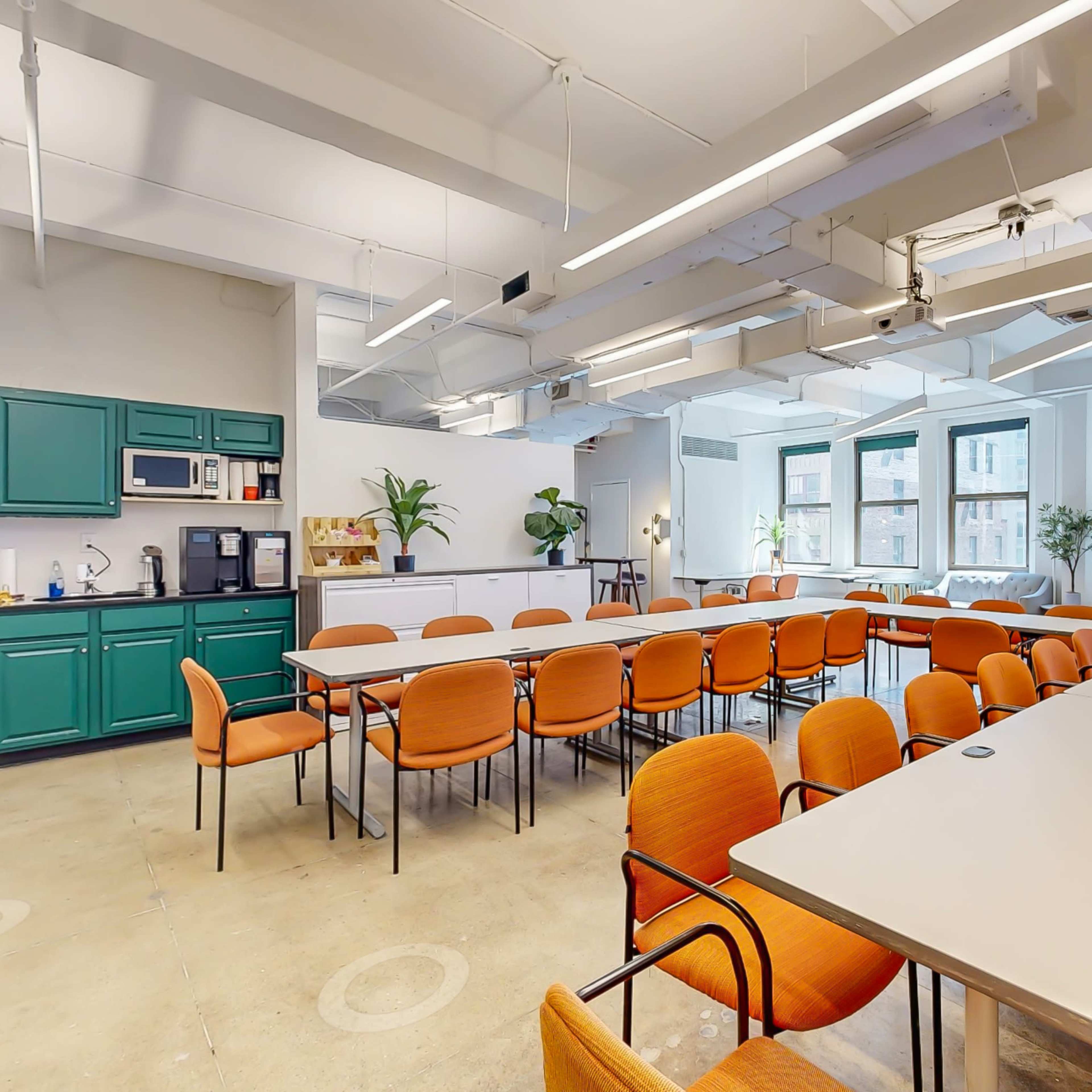 A modern conference room features long tables with orange chairs, green cabinetry, and large windows allowing natural light.