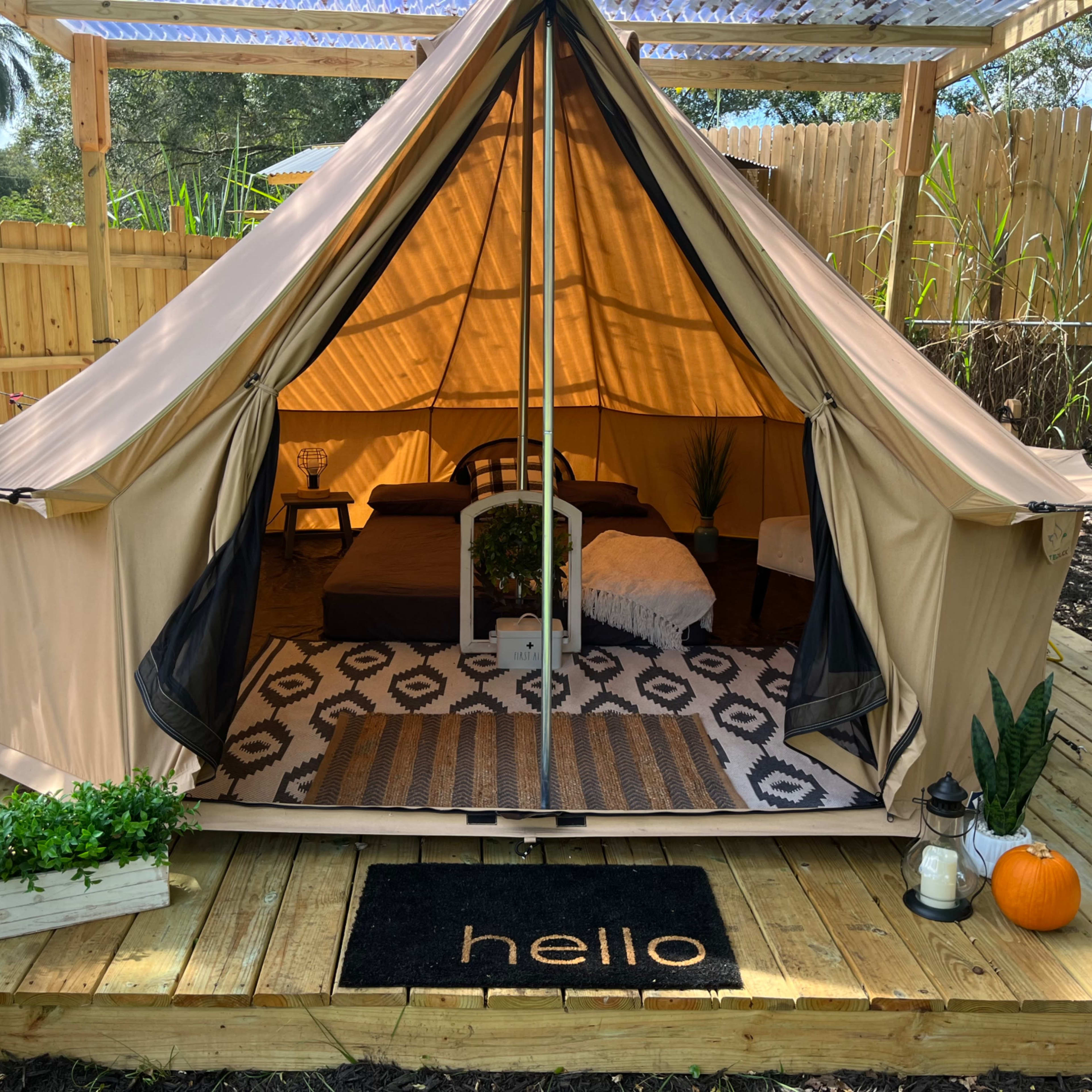 A beige tent set up on a wooden deck, featuring a bed, a decorative rug, a potted plant, and a welcome mat that reads "hello."