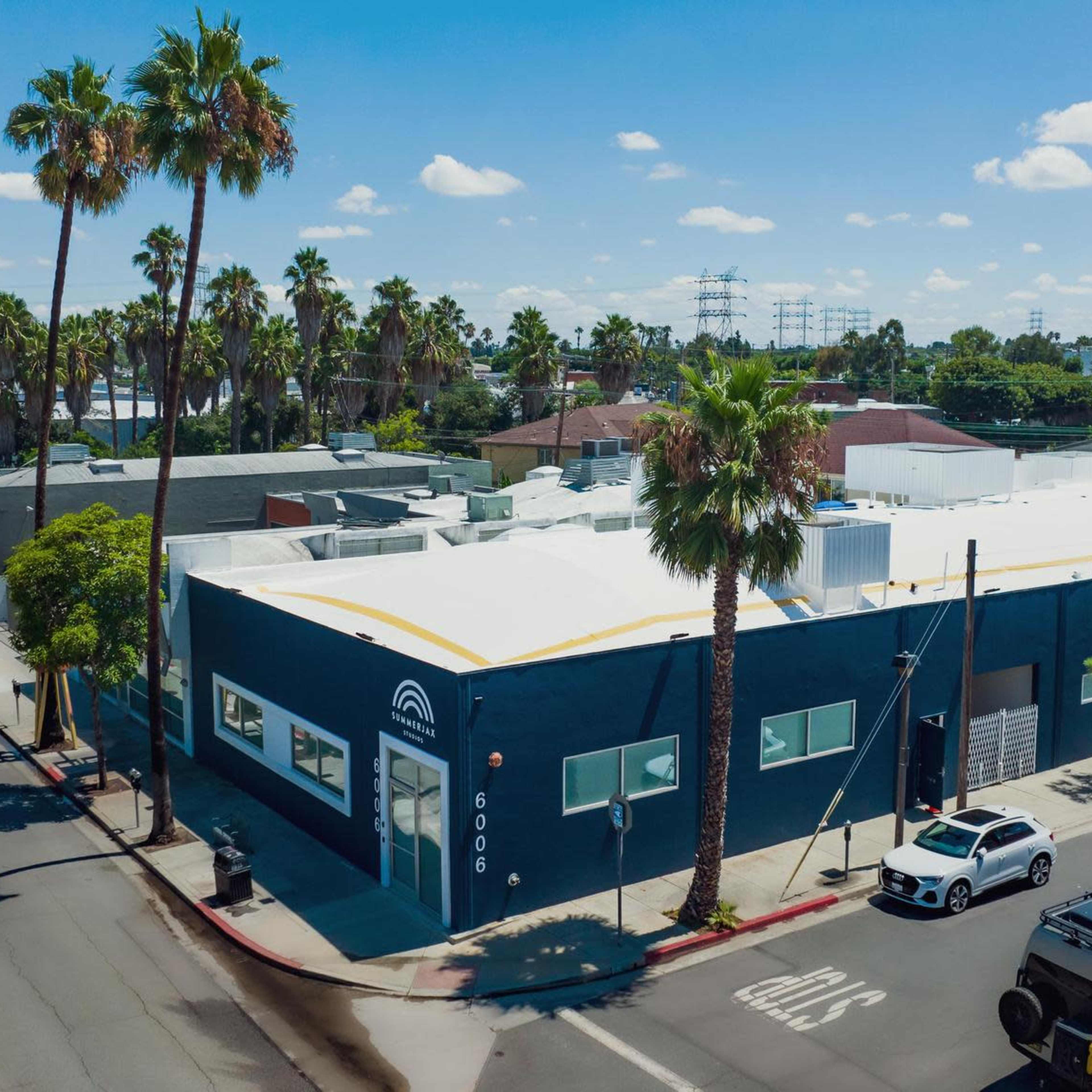 A blue commercial building with a white roof sits at the corner of a street lined with palm trees, surrounded by other low-rise structures and a tall building in the background.