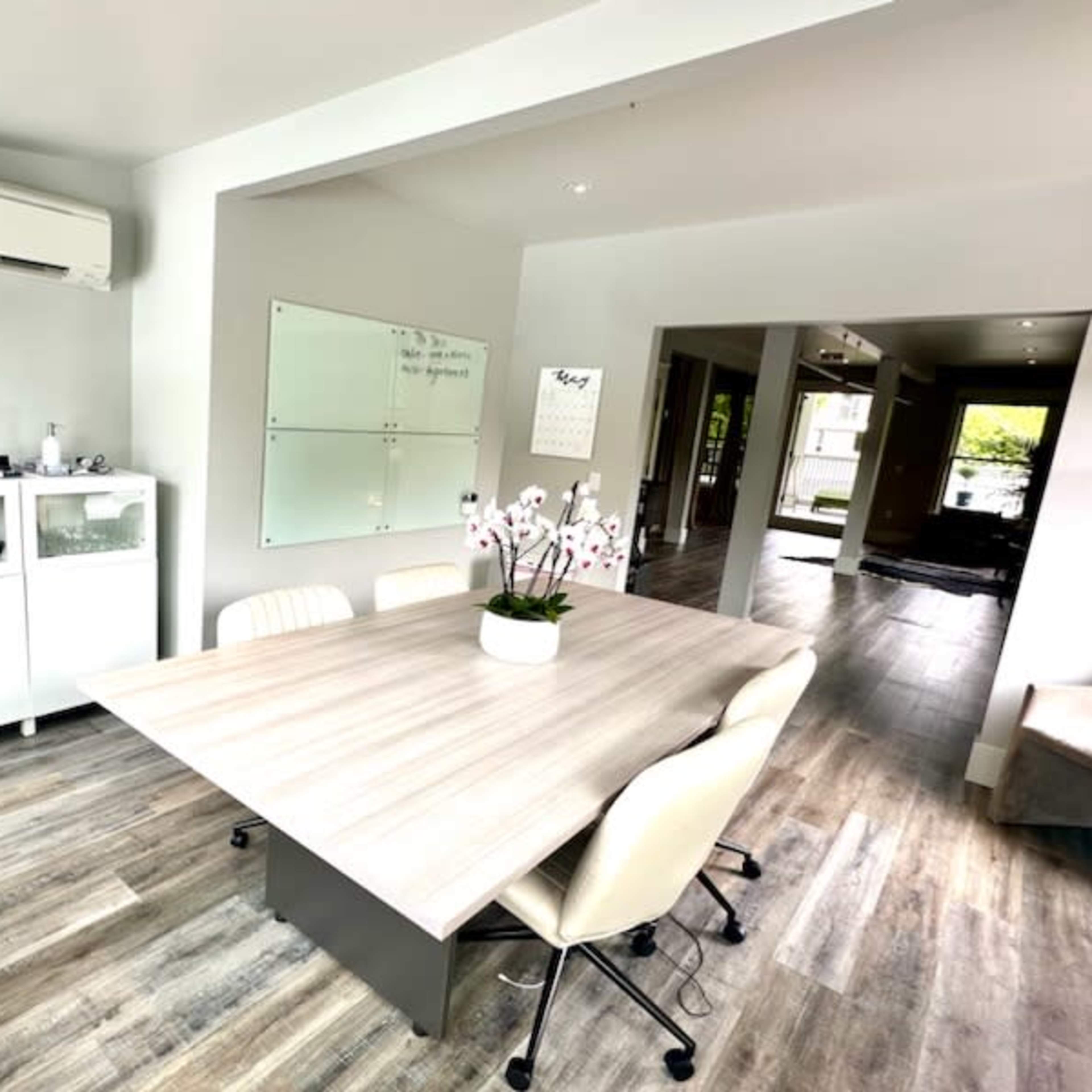 A modern conference room features a wooden table surrounded by beige chairs, with a glass whiteboard and decorative plants in the space.