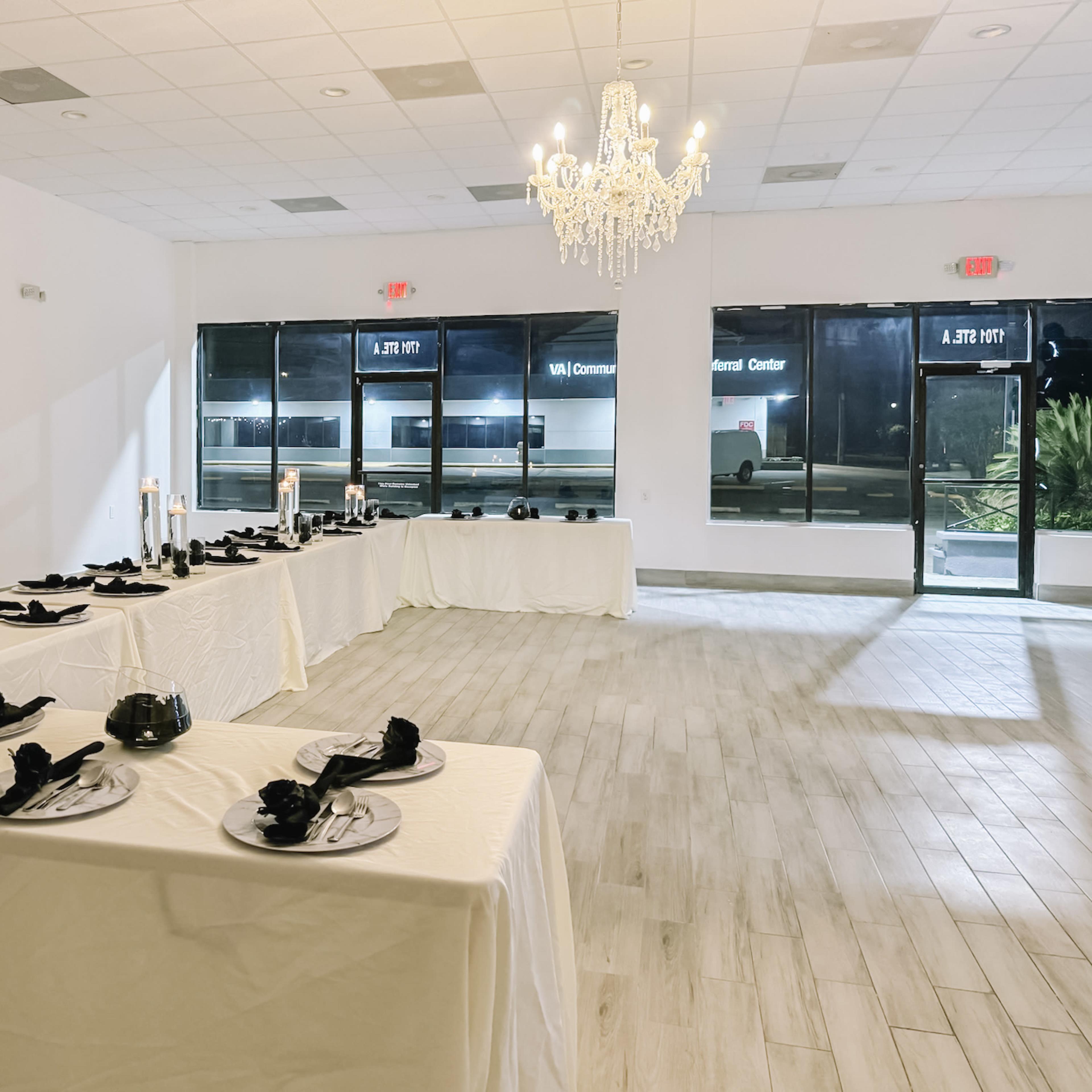 A banquet hall with two long tables covered in white cloths, set with black napkins and plates, beneath a chandelier.