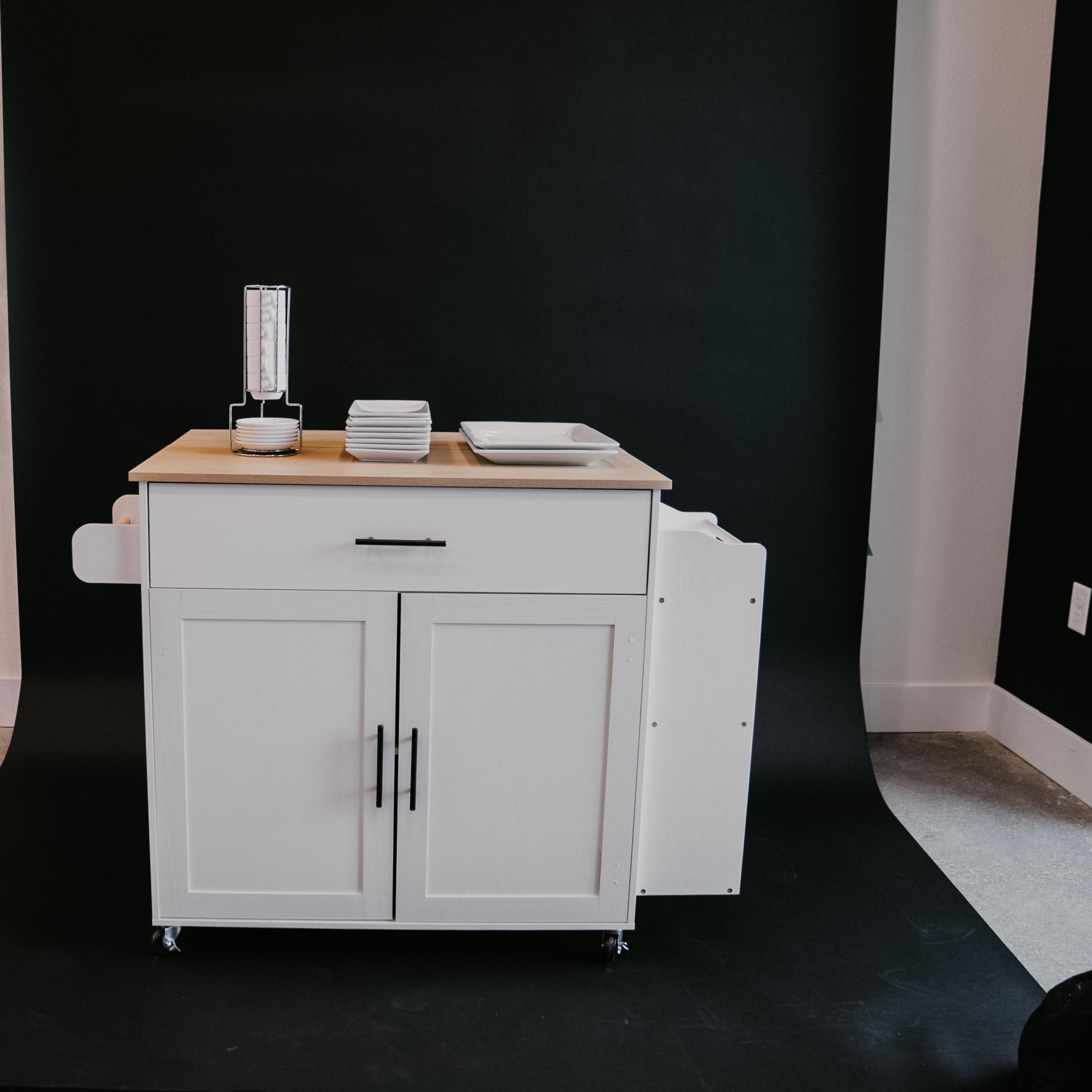 A white kitchen cart with two drawers and a wooden top is placed against a black backdrop, accompanied by plates and a table lamp.