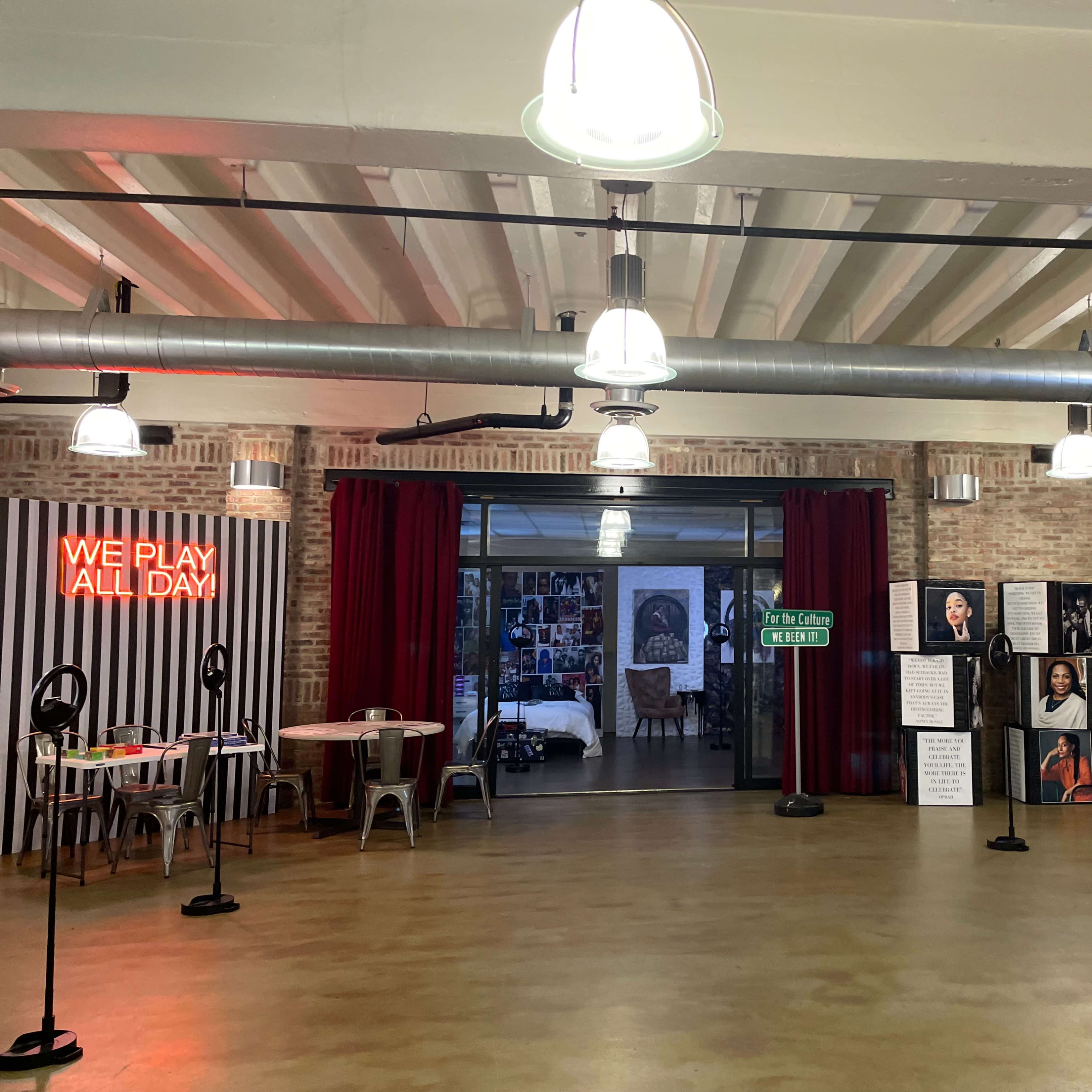 A spacious, modern room with a neon sign reading "WE PLAY ALL DAY," featuring brick walls, poster displays, and several tables and chairs.