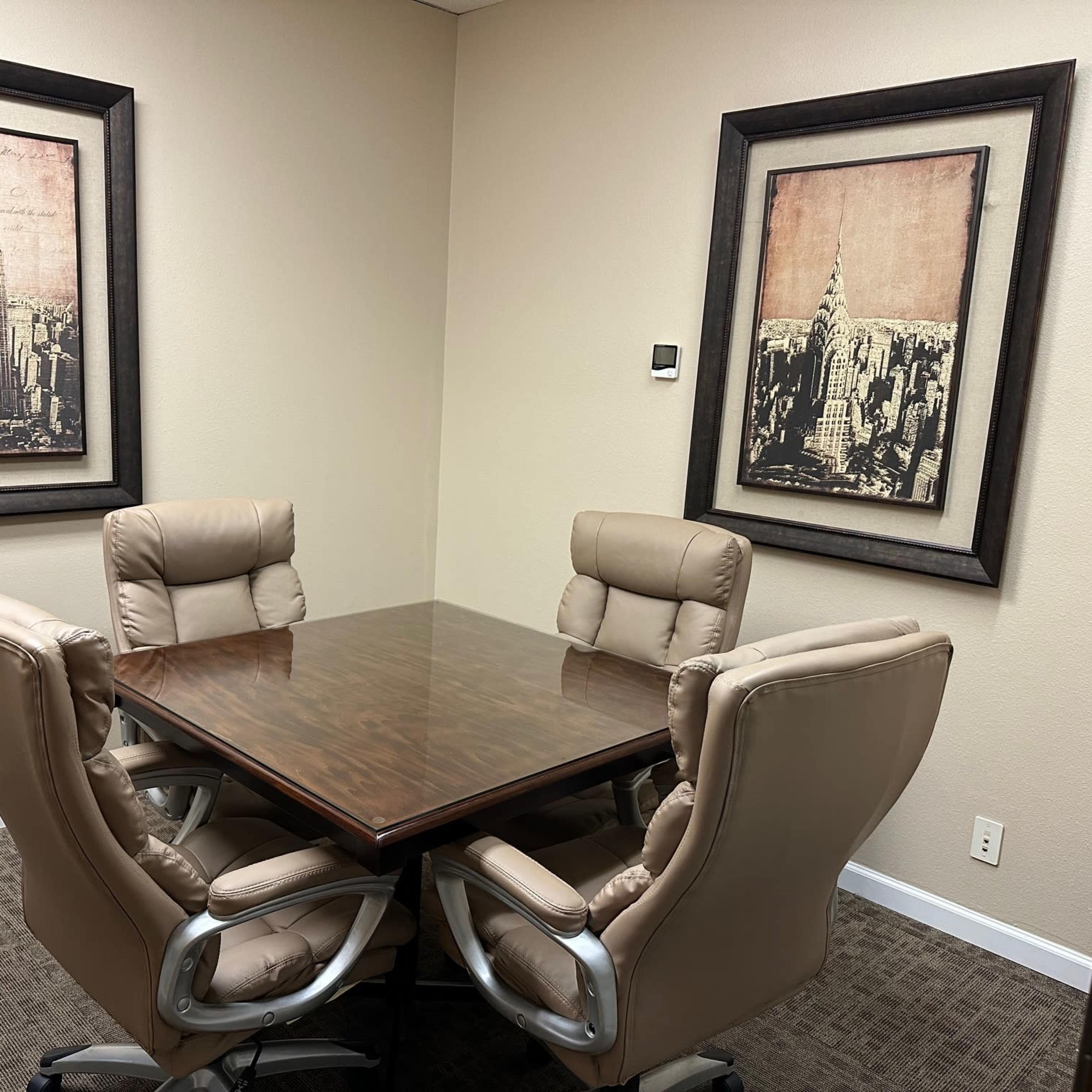 A small conference room with a wooden table surrounded by four beige executive chairs, and two framed cityscape artworks on the wall.